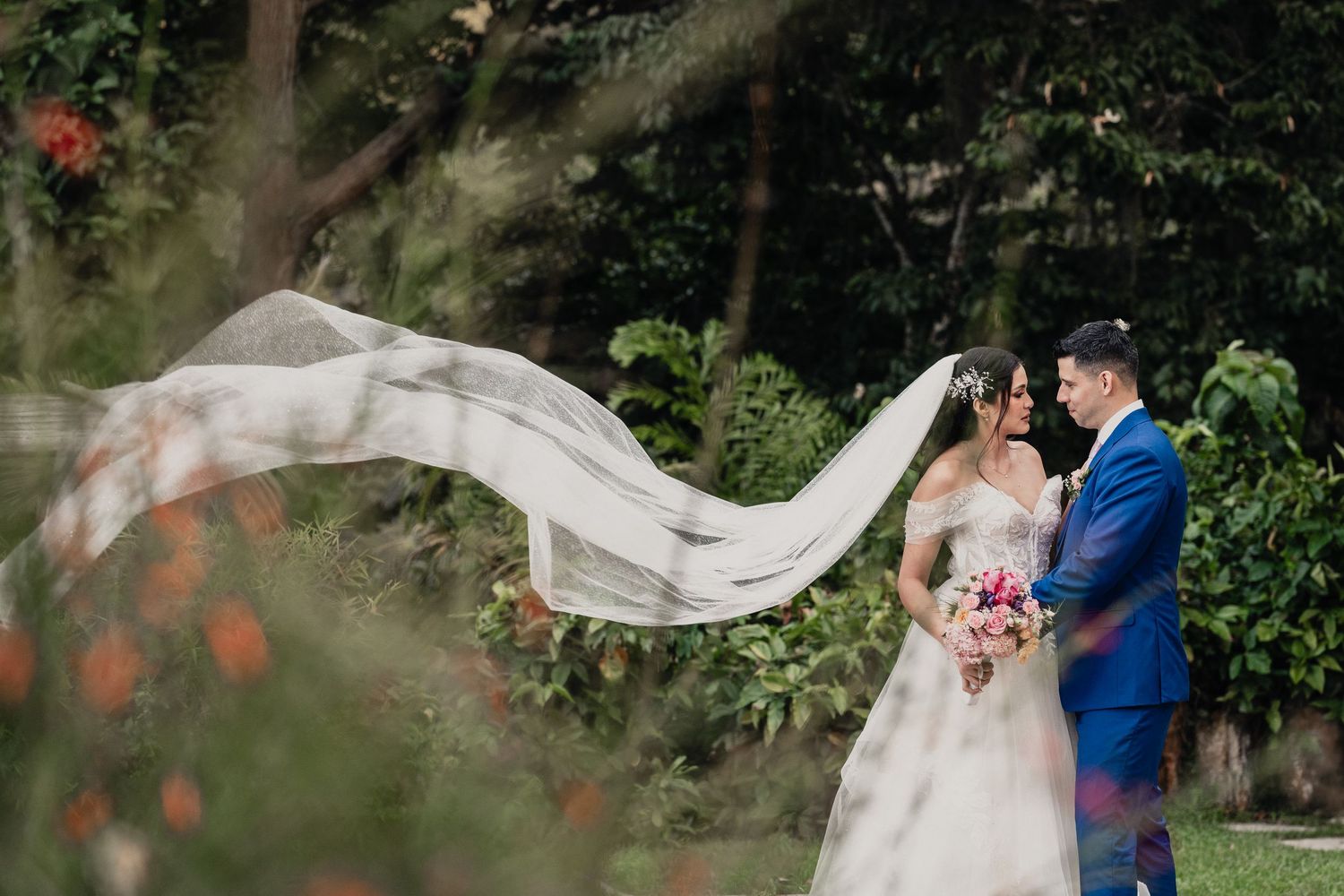 A bride's veil flows dramatically in the wind during an outdoor wedding photoshoot.