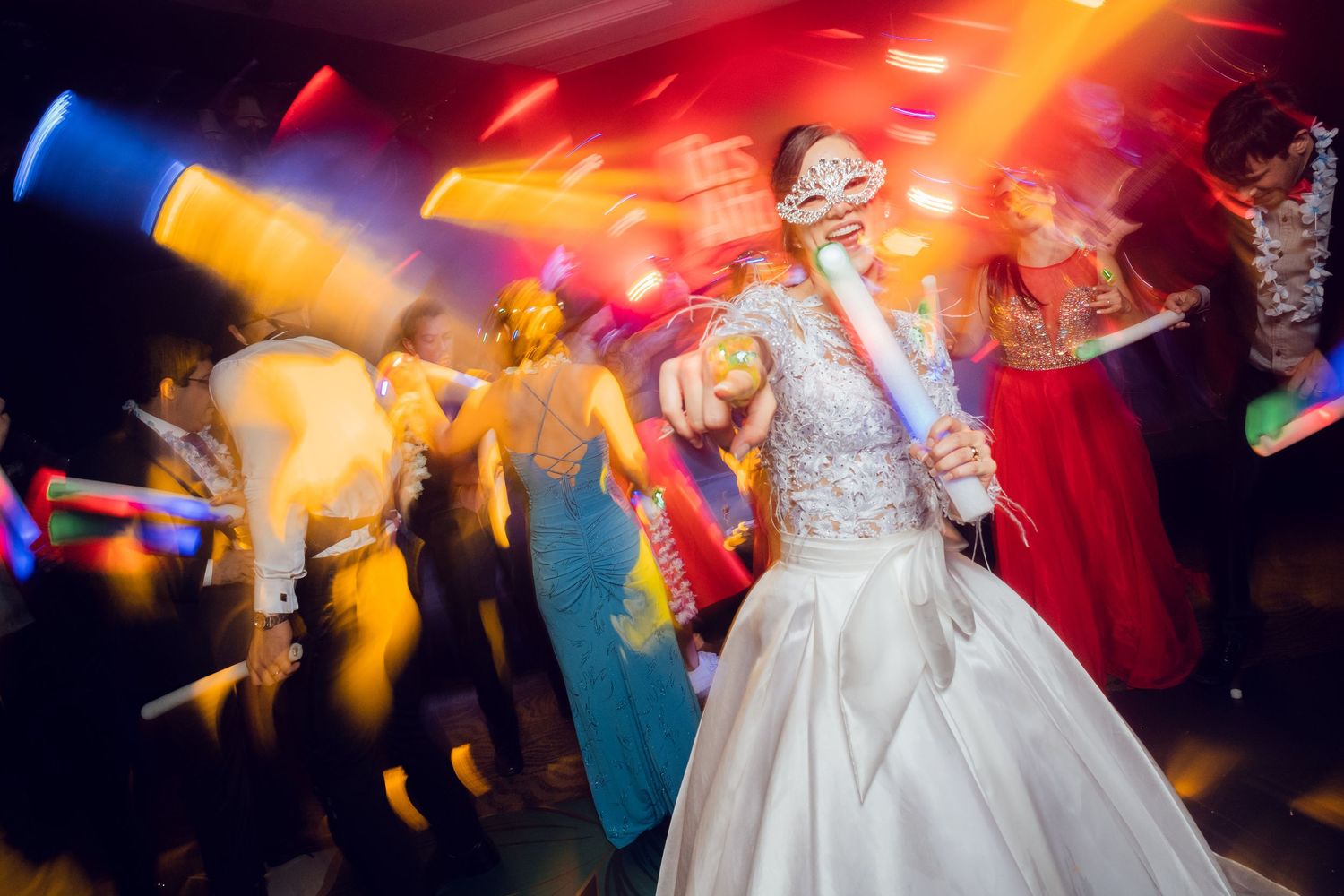 Wedding reception dance party with people dancing under colorful disco lights as someone in a white wedding gown leads the celebration.
