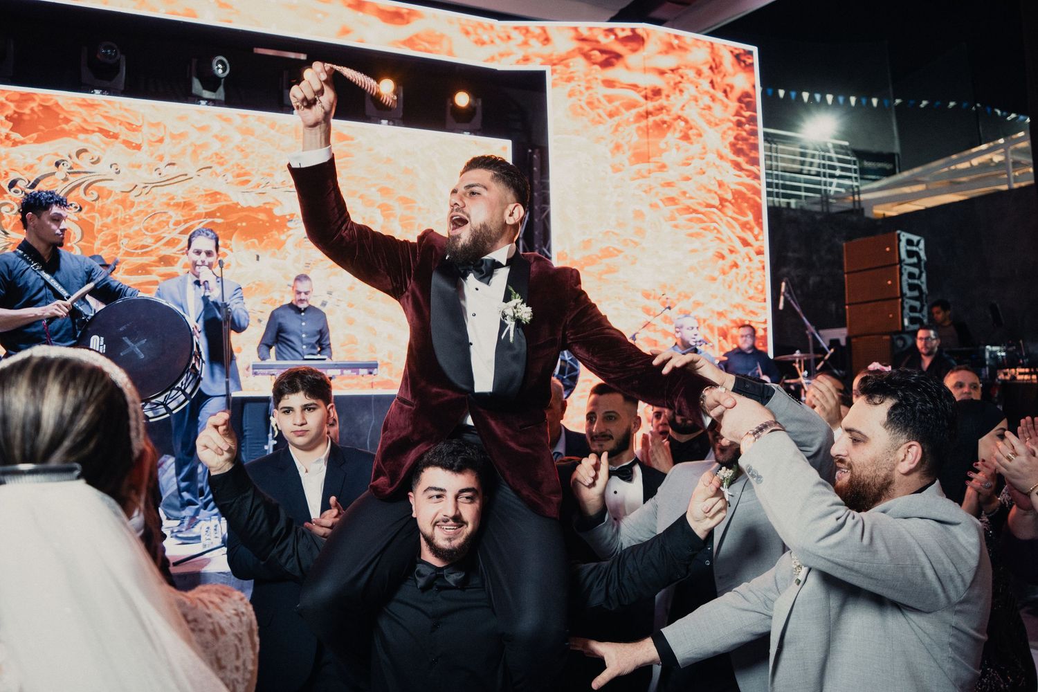 Wedding celebration showing guests lifting and cheering in front of an orange-lit backdrop.