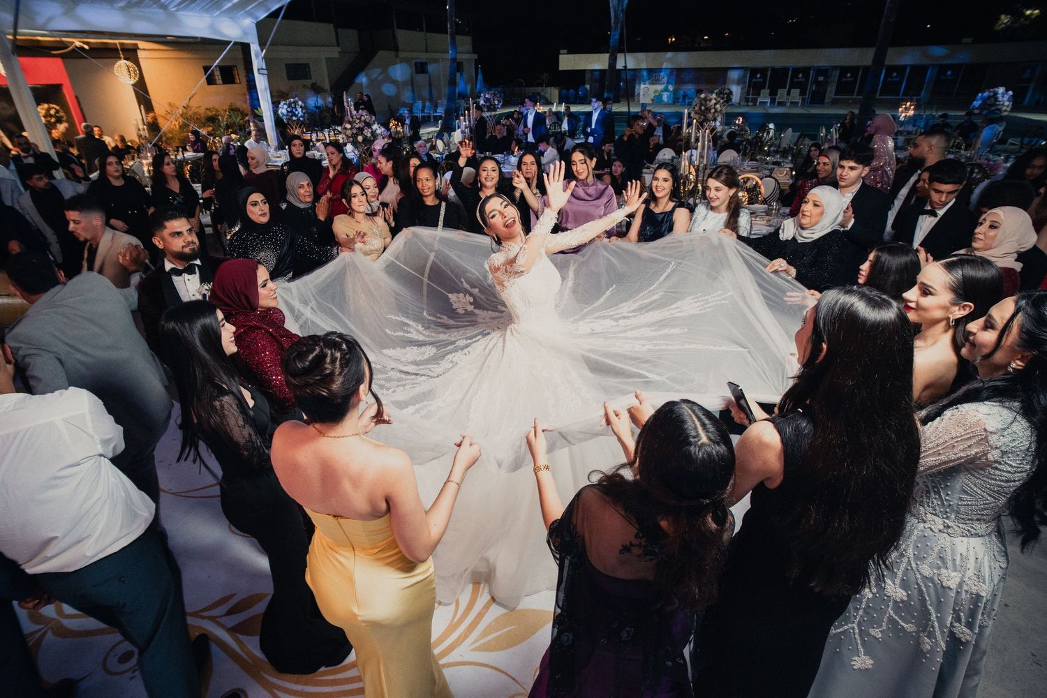 Wedding guests gather in a circle on the dance floor, lifting the bride's veil in celebration.