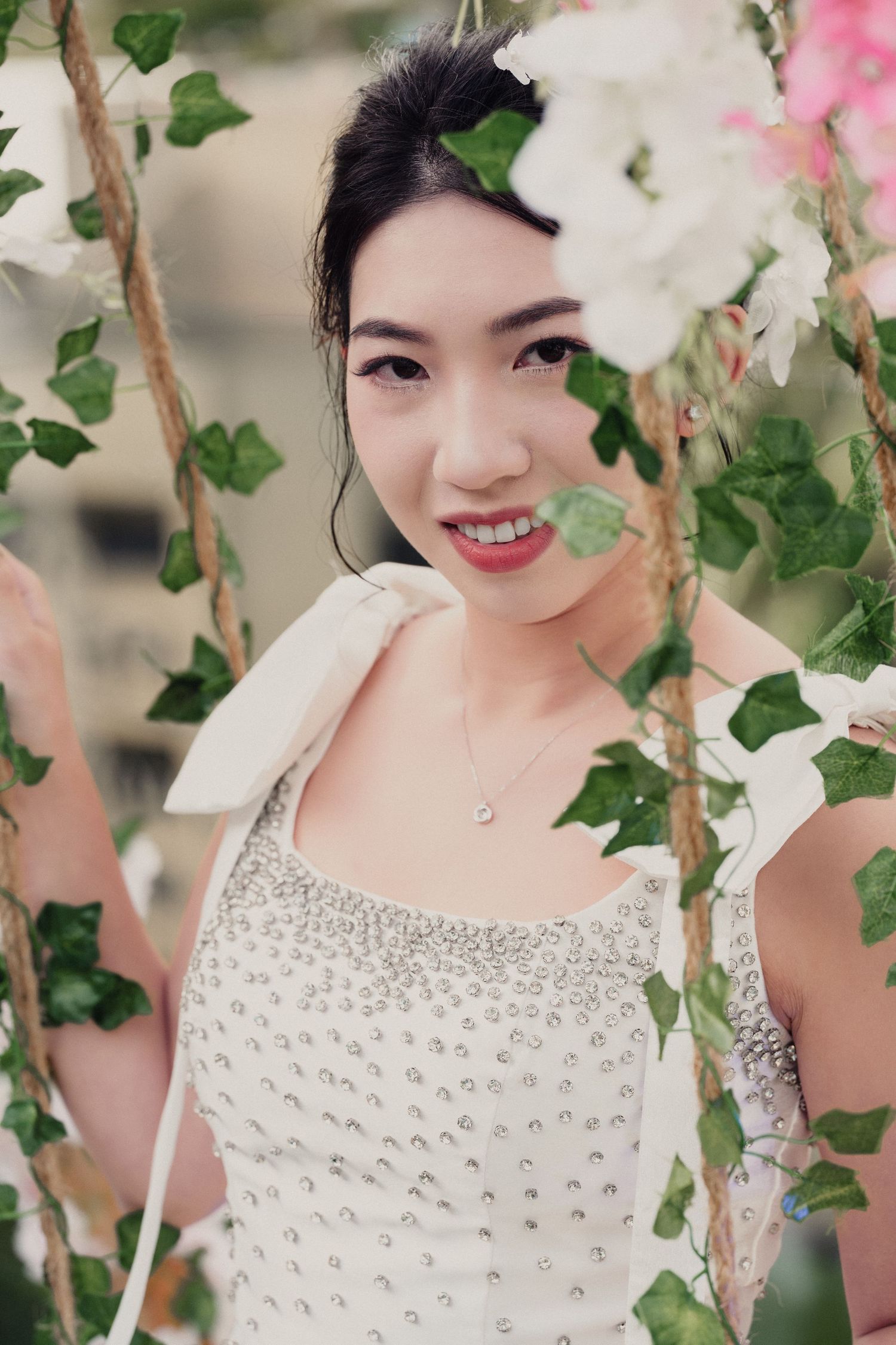 A person in a white beaded dress poses among green ivy and pink flowers in a romantic garden setting.