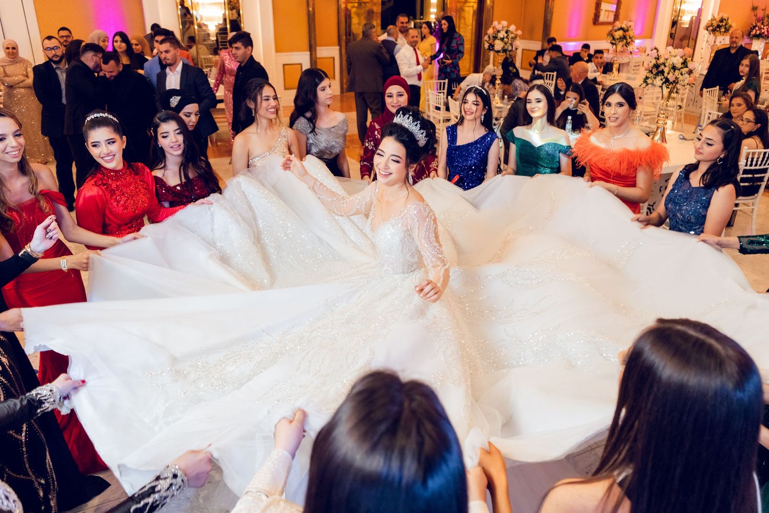 A bride in a flowing white ballgown twirls on the dance floor surrounded by wedding guests at an elegant reception.