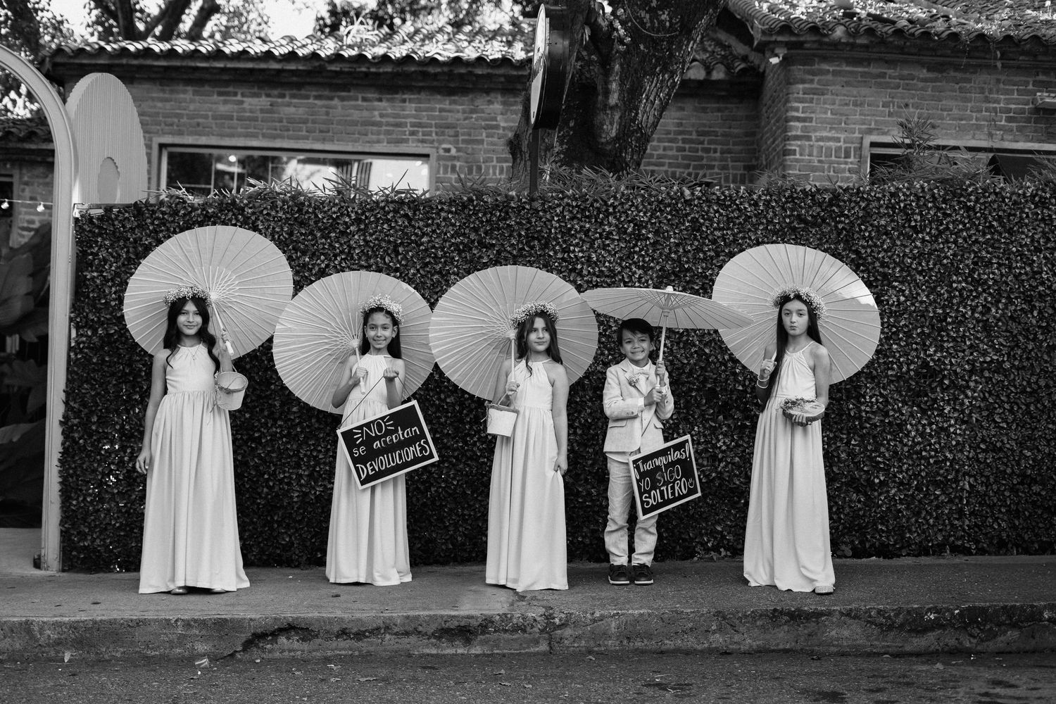 A row of flower girls in white dresses standing against a brick wall with umbrellas.