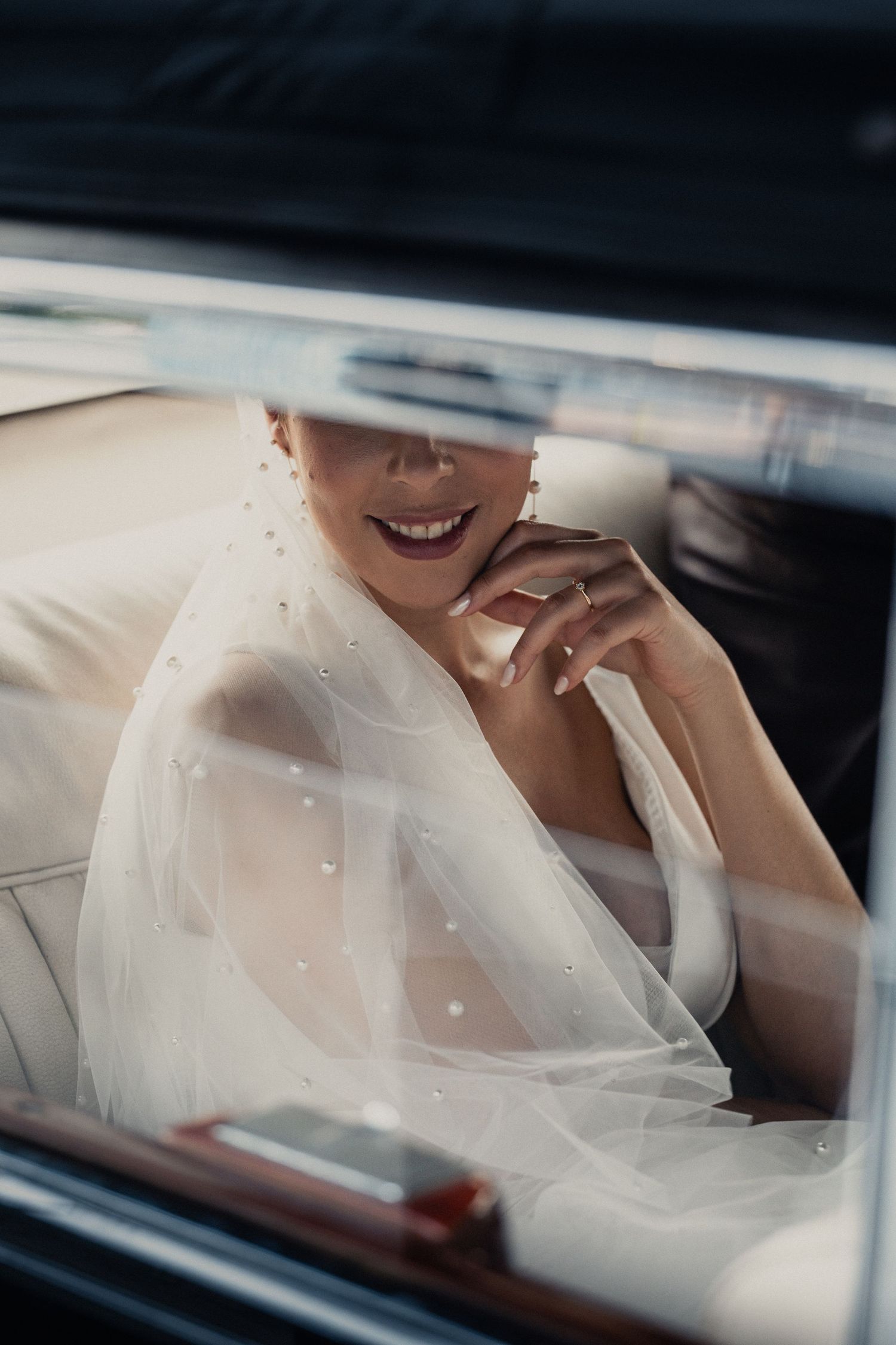 A joyful bride in white veil sits in a vintage car, her bright smile visible through delicate tulle fabric.