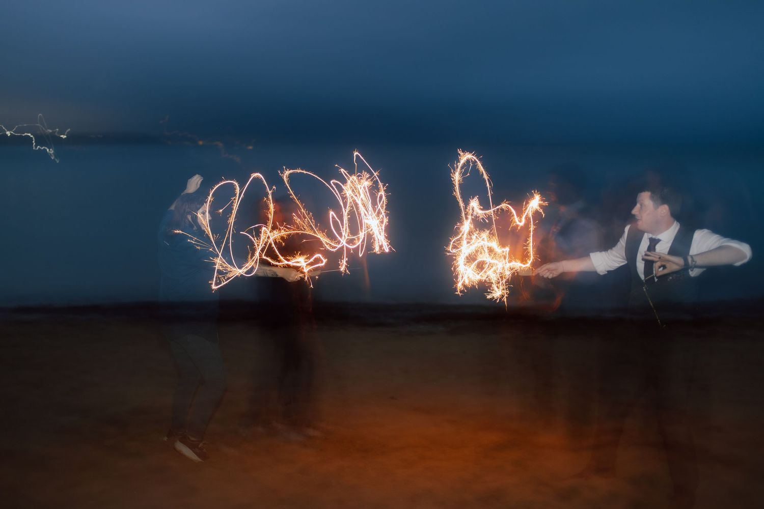 Long exposure photograph of light painting with sparklers creating glowing trails on a dark beach at night.