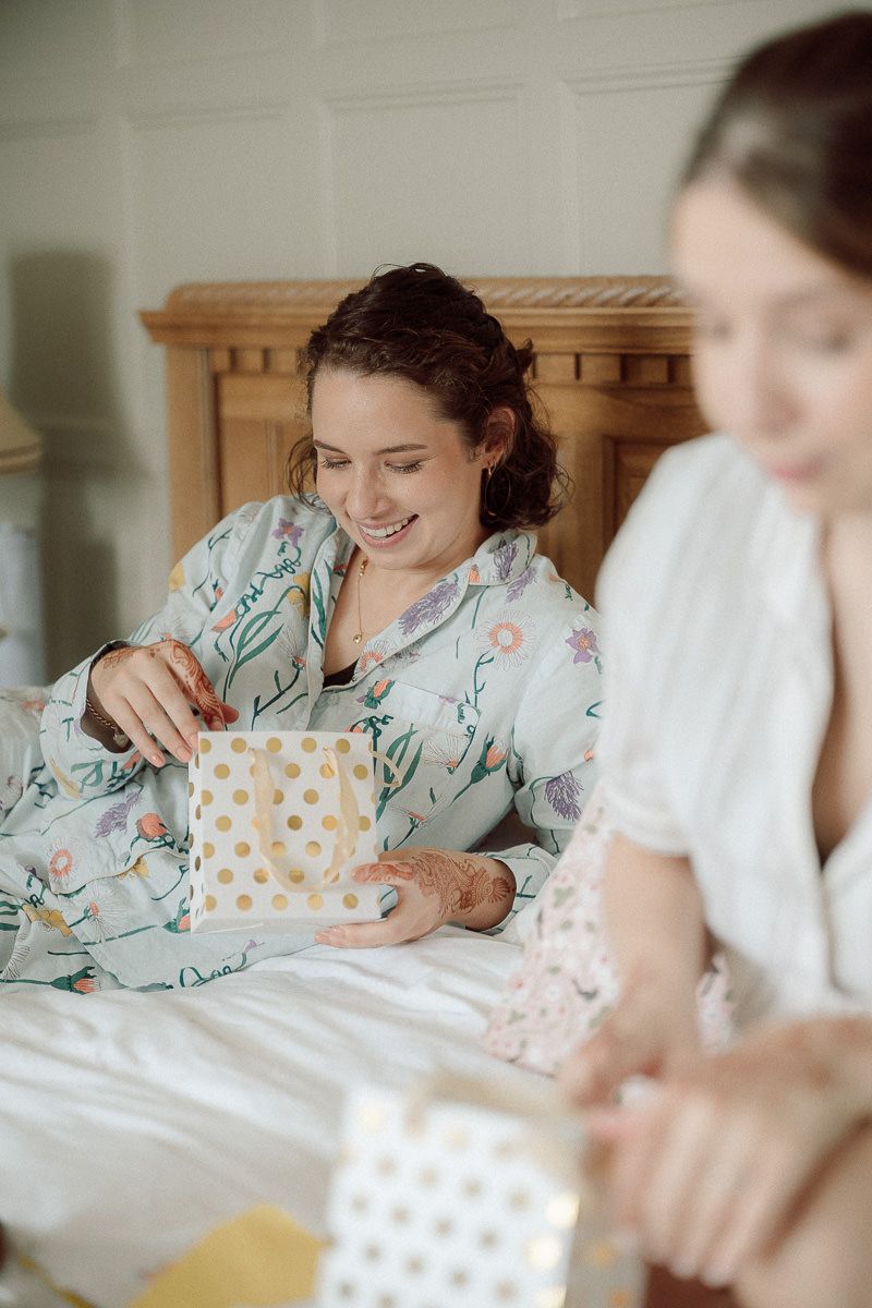 Two friends in sleepwear sit on a bed opening polka dot gift boxes during a cozy morning gathering.