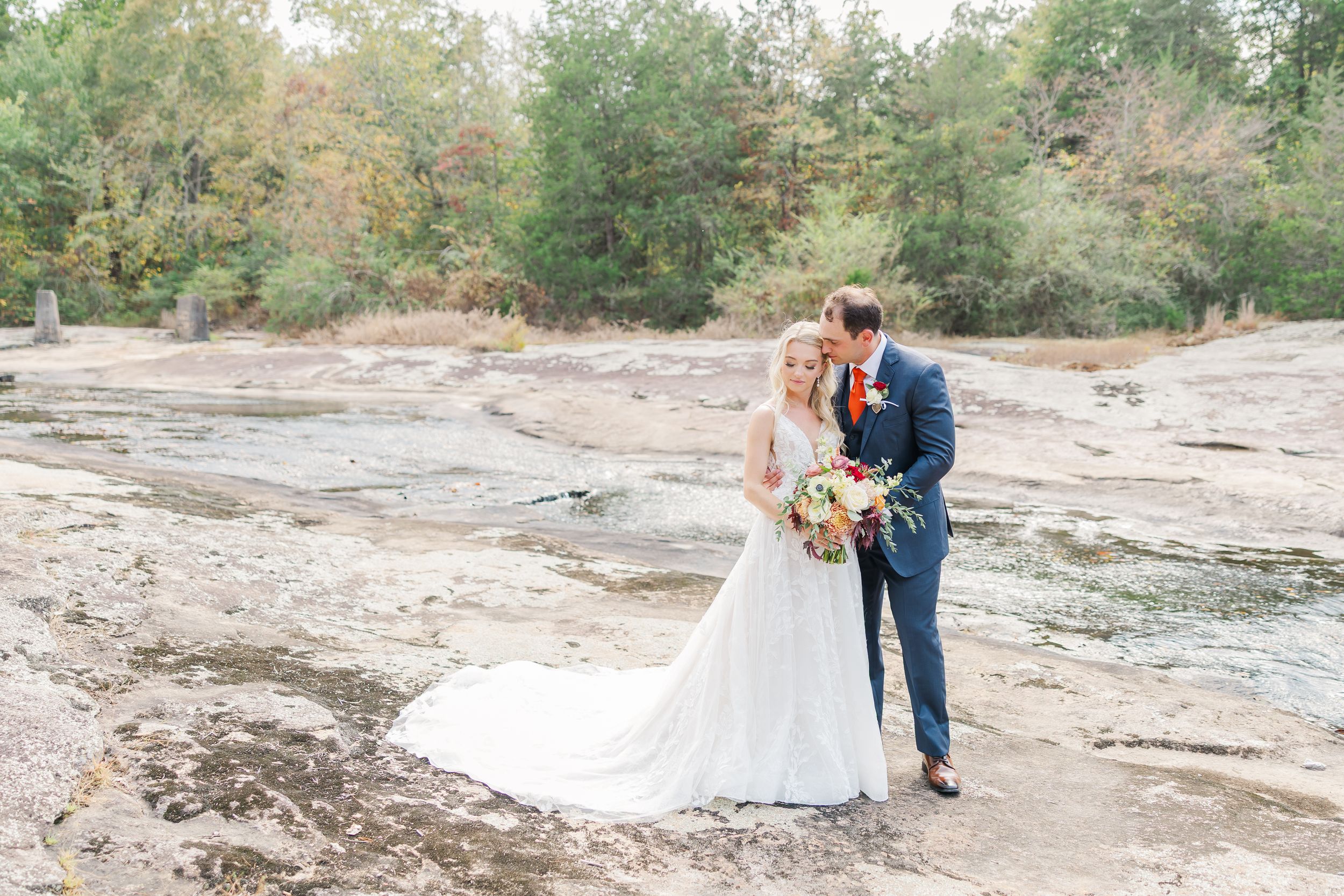 A couple in wedding attire shares a romantic moment while walking along a rocky creek bed surrounded by autumn trees at Mill at Fine Creek