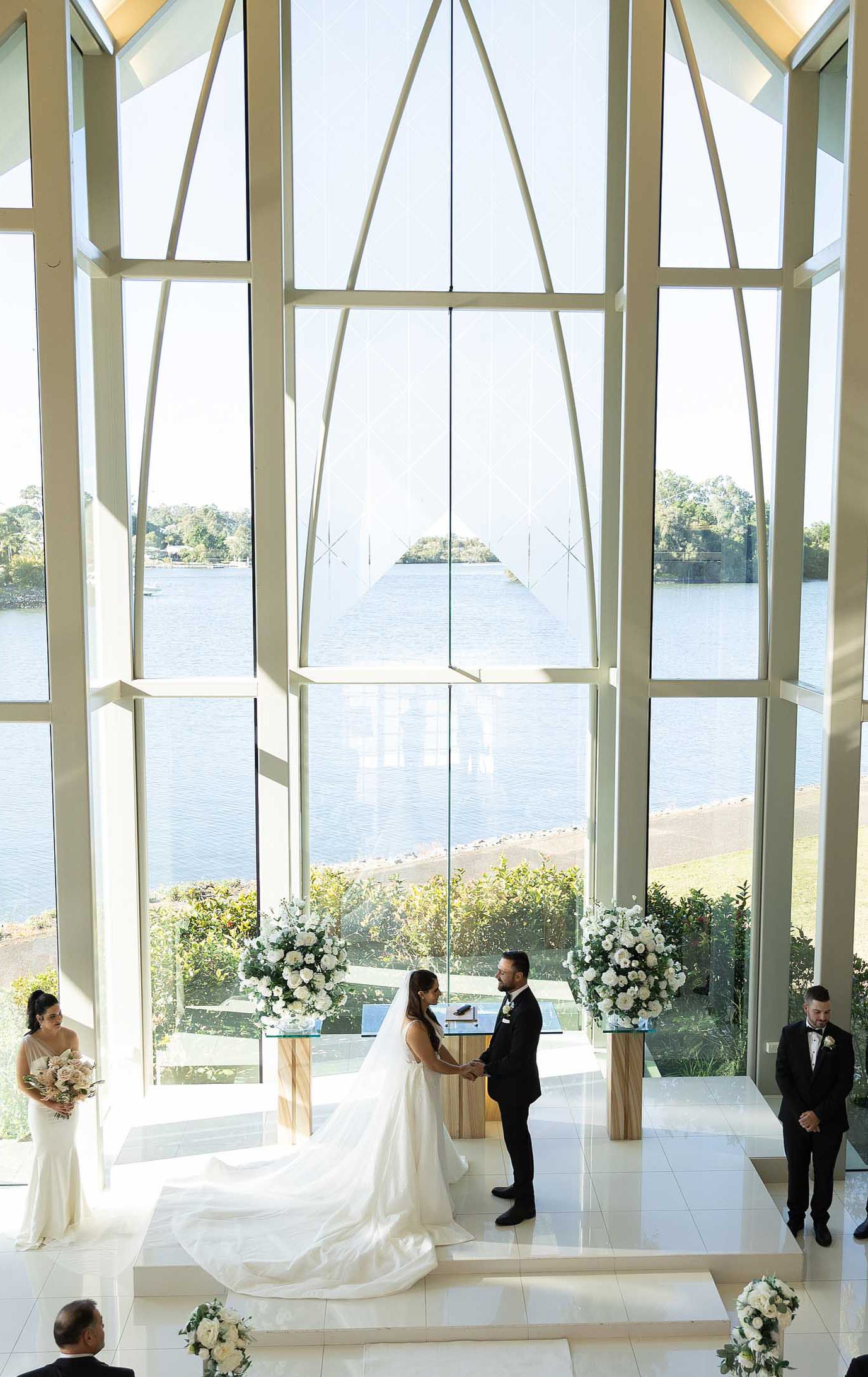 Bride and groom exchange vows in a breathtaking glass chapel with panoramic waterway views and white florals.