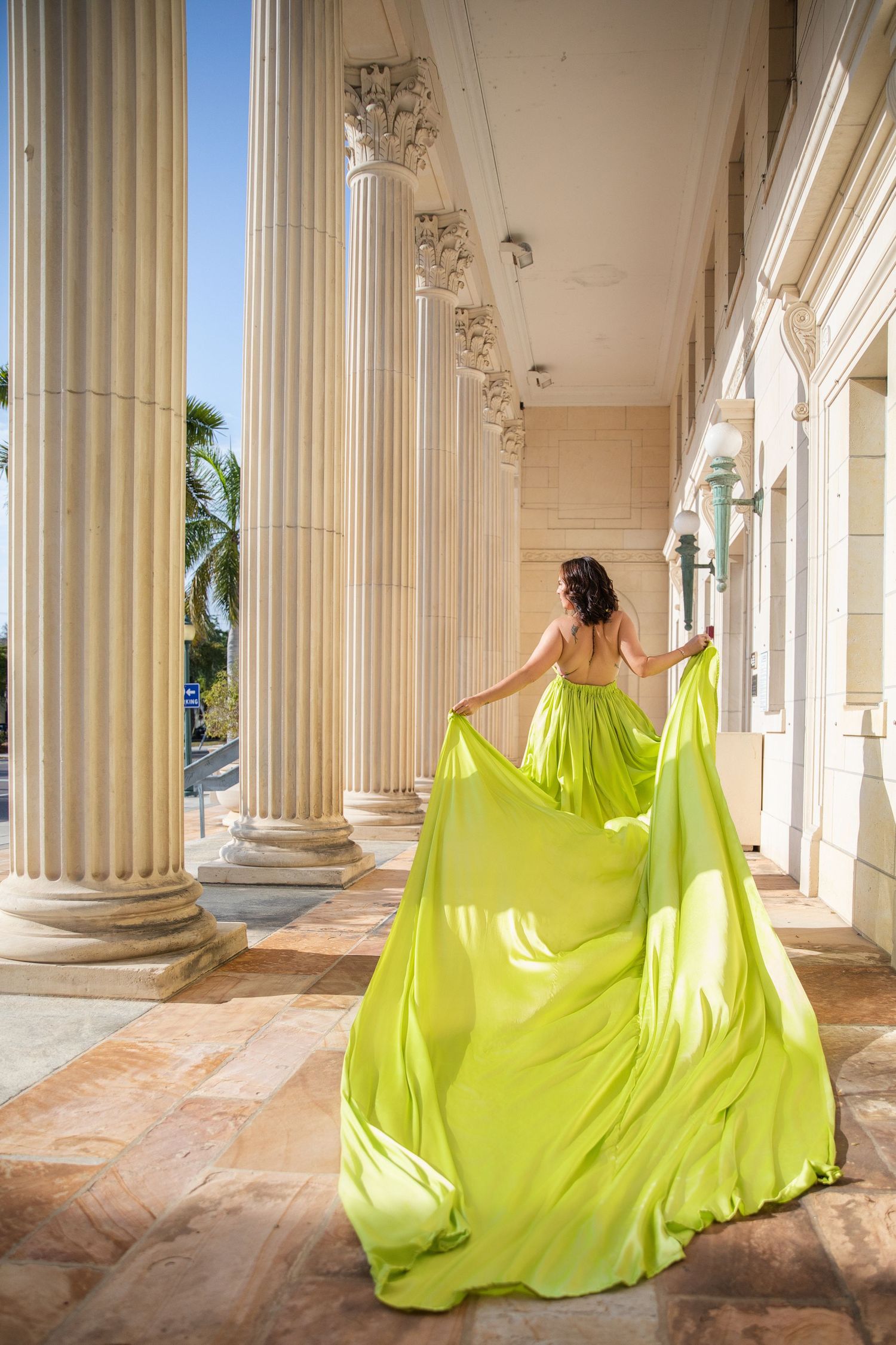 A figure in a flowing neon yellow gown moves gracefully through a classical colonnade with white pillars.