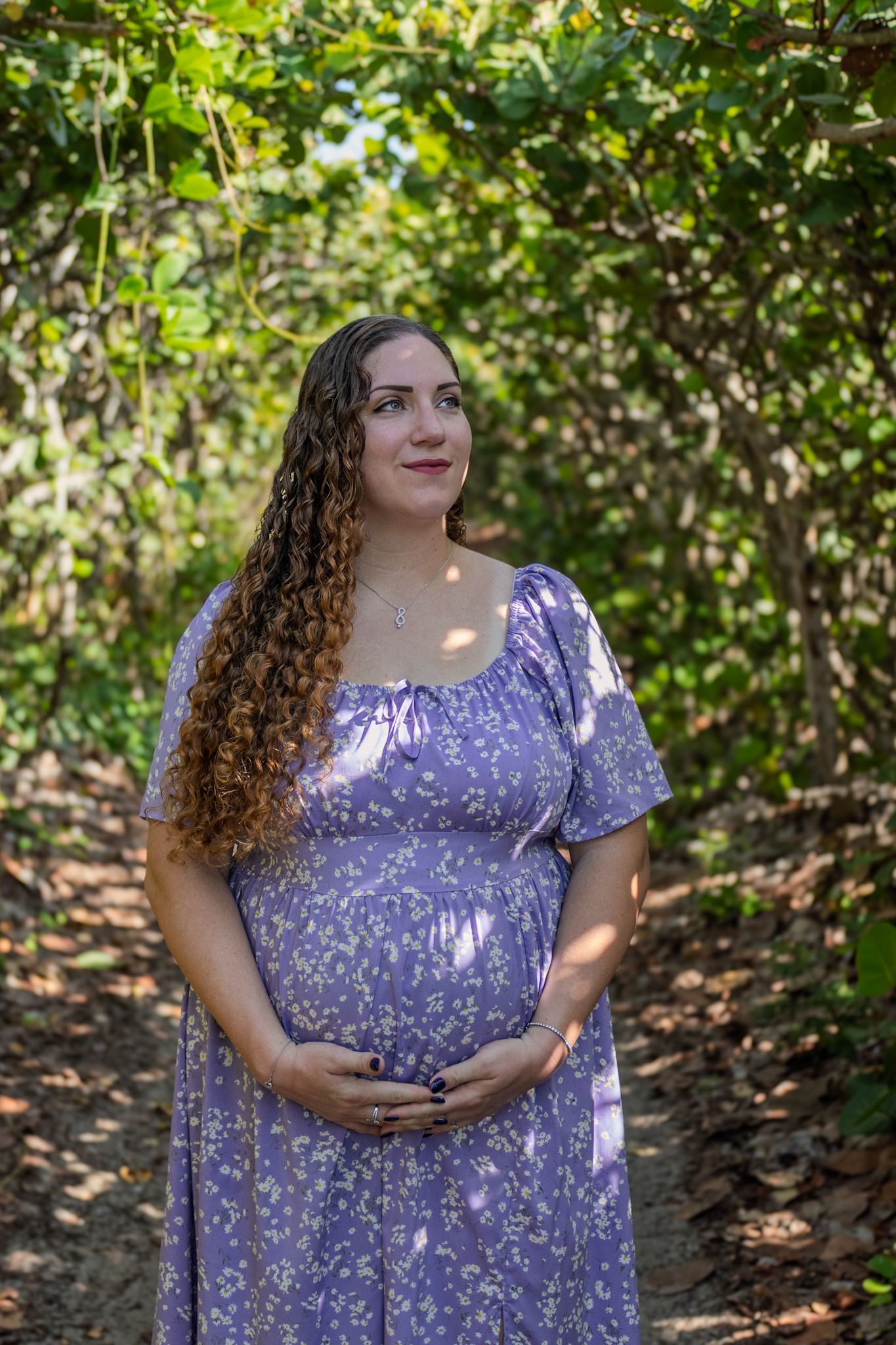 A person in a purple floral dress stands in dappled sunlight among trees and foliage in a natural outdoor setting.