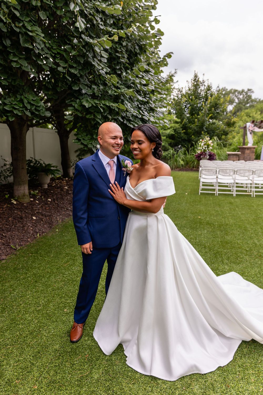 Elegant wedding couple posing on a manicured lawn with white chairs in the background.