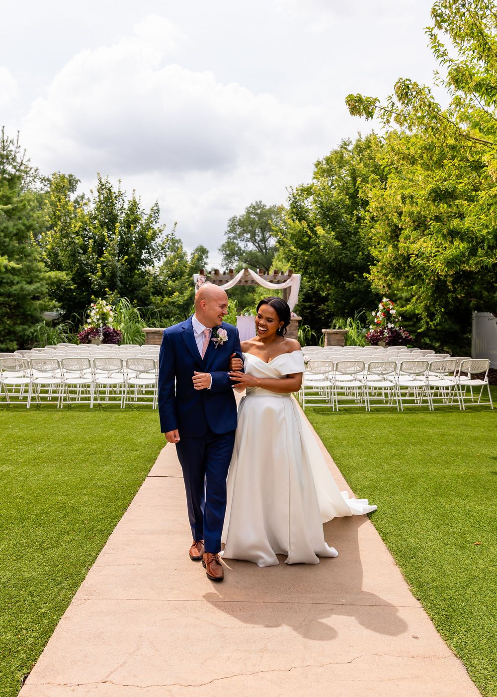 Newlyweds walking along a stone pathway with white ceremony chairs visible in the garden at Villa Aletta in Omaha, NE.