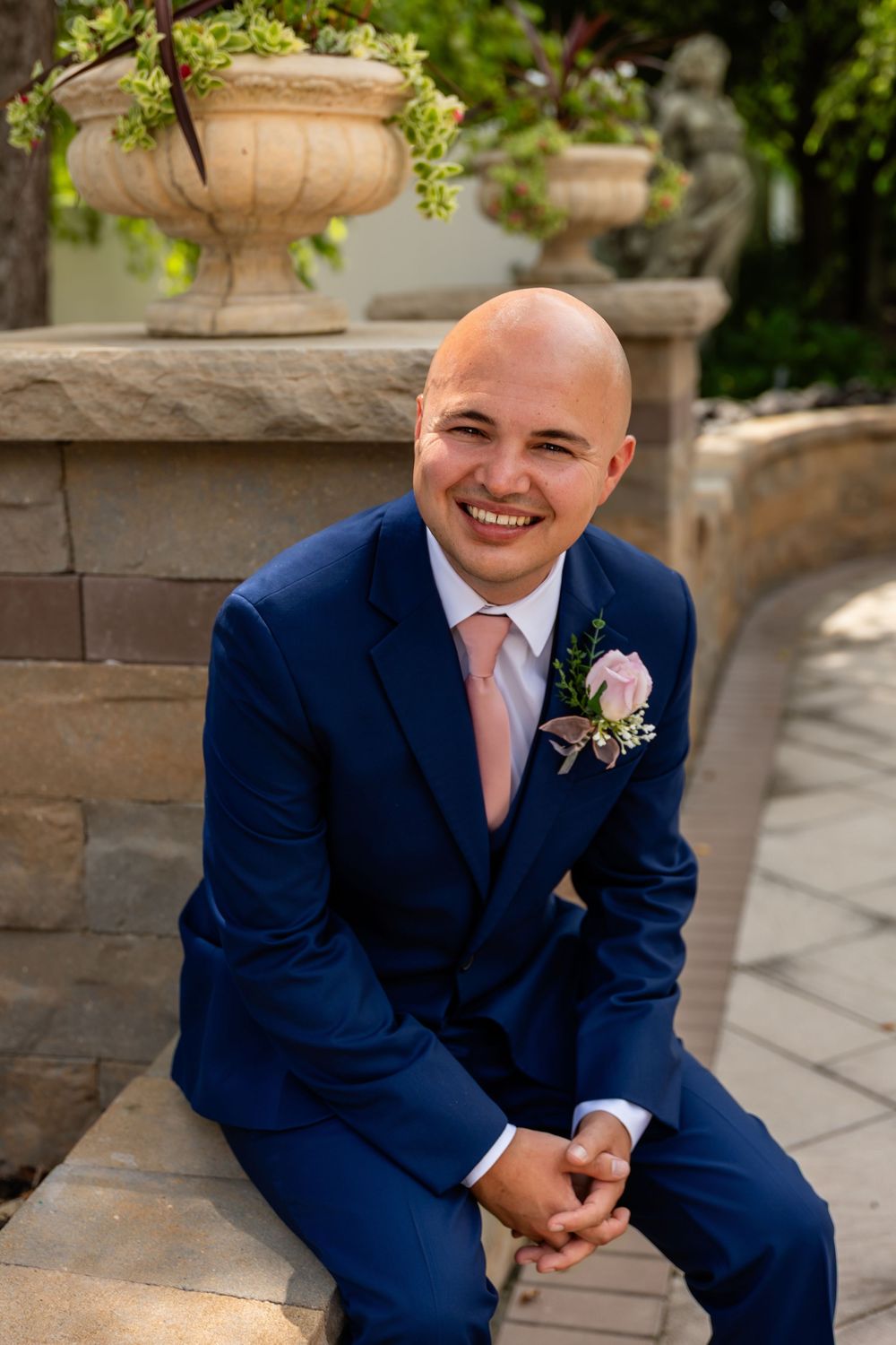 Groom in navy suit with pink boutonniere smiling in an outdoor garden setting.