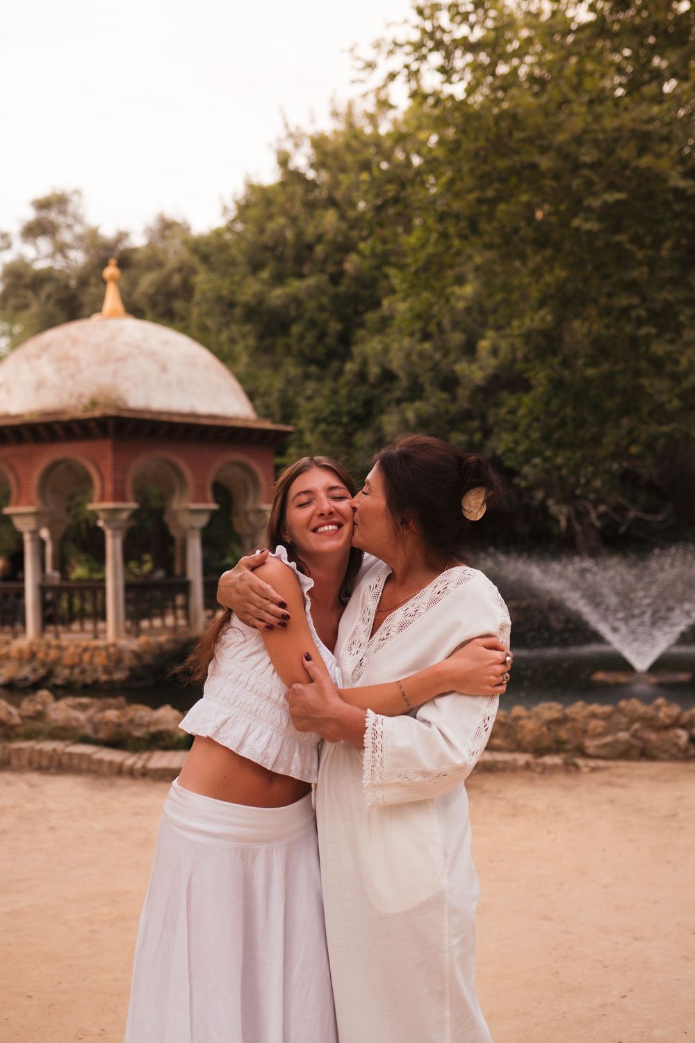 Two women in white outfits embrace and share a kiss near a fountain and ornate gazebo outdoors.