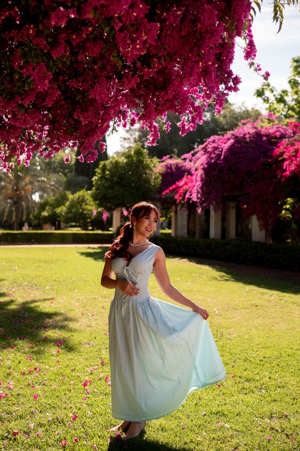 Woman in light blue dress twirling under vibrant pink bougainvillea trees in a lush green garden.