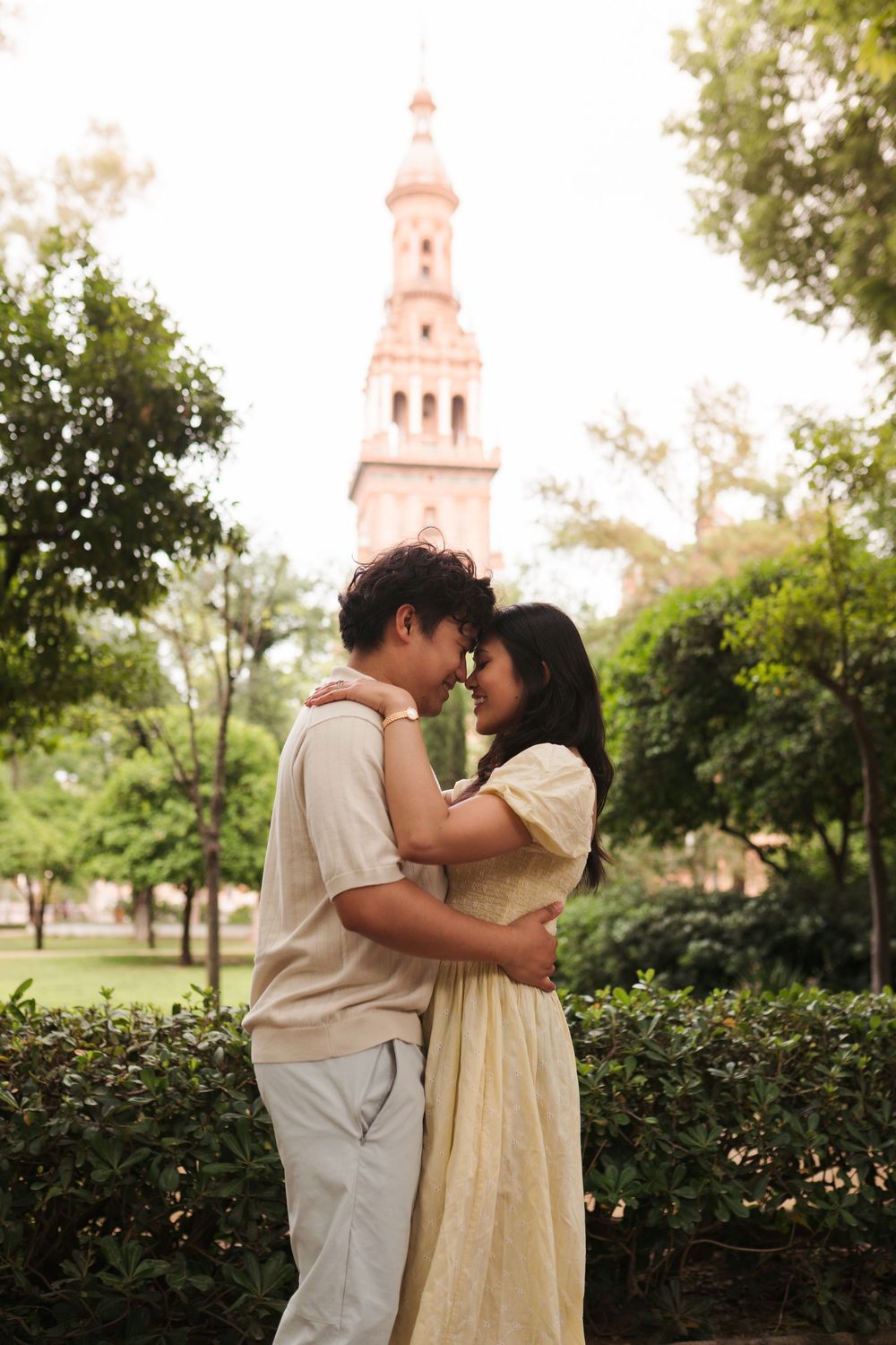 Romantic couple embracing in a lush green park with a historic tower in the background.