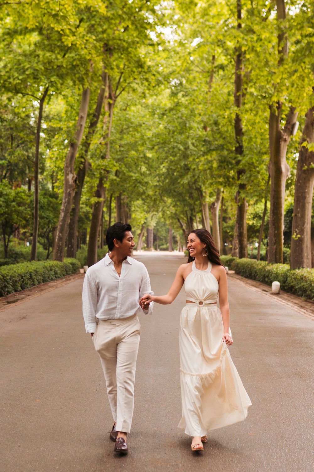 Happy couple holding hands walking down a tree-lined park path, woman in cream dress, man in white outfit.