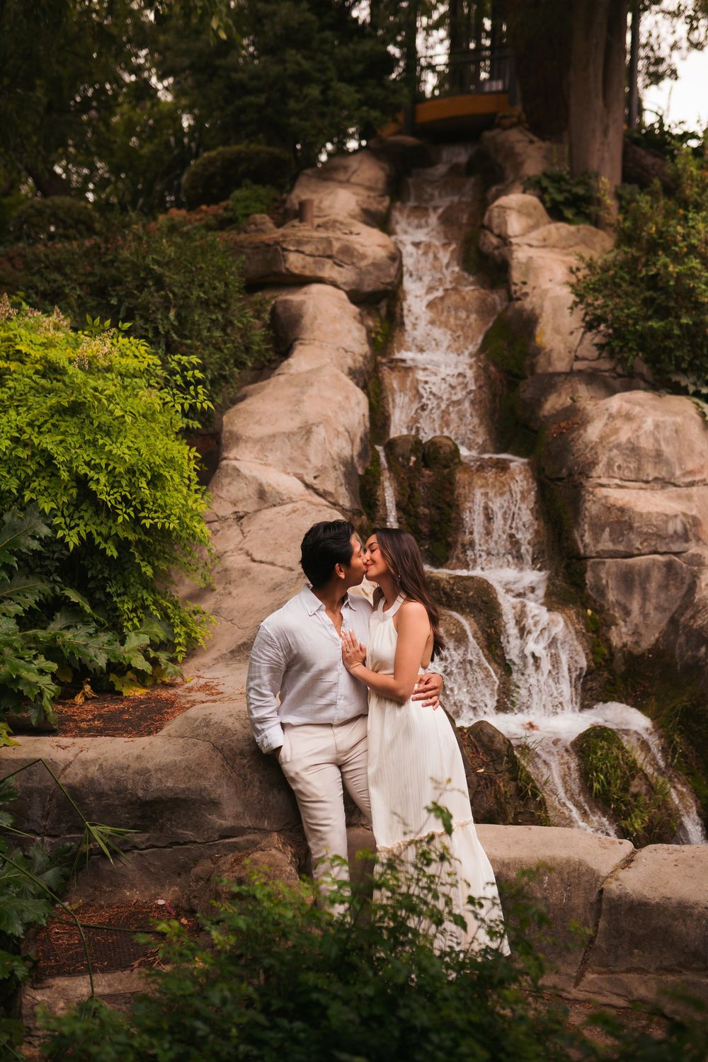 Couple sharing a romantic kiss in front of a cascading waterfall surrounded by lush greenery.