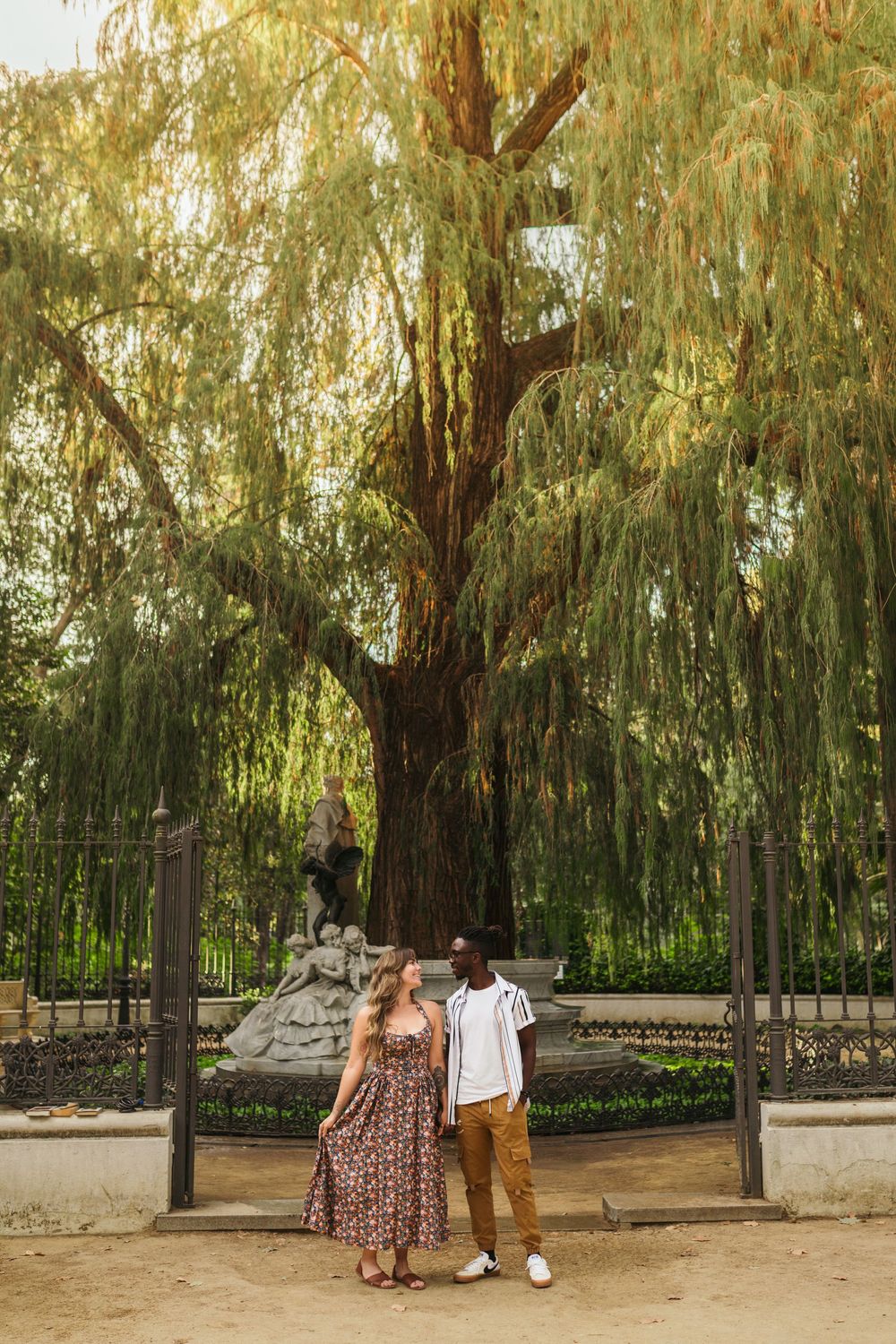 A couple stands beneath a majestic weeping willow tree in a peaceful garden setting.