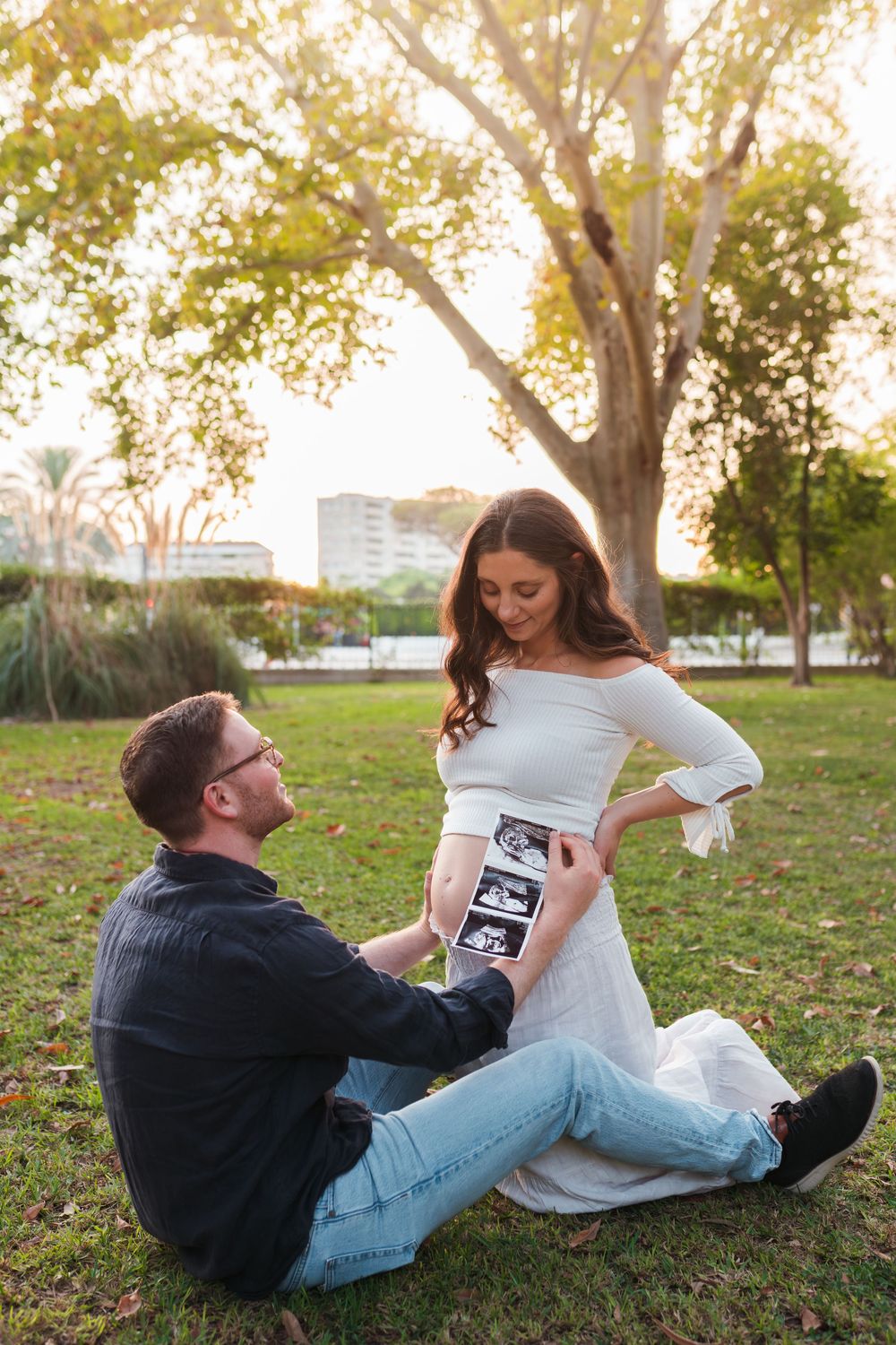 A playful couple rolls around on grass while looking at an ultrasound photo during a pregnancy announcement photoshoot.