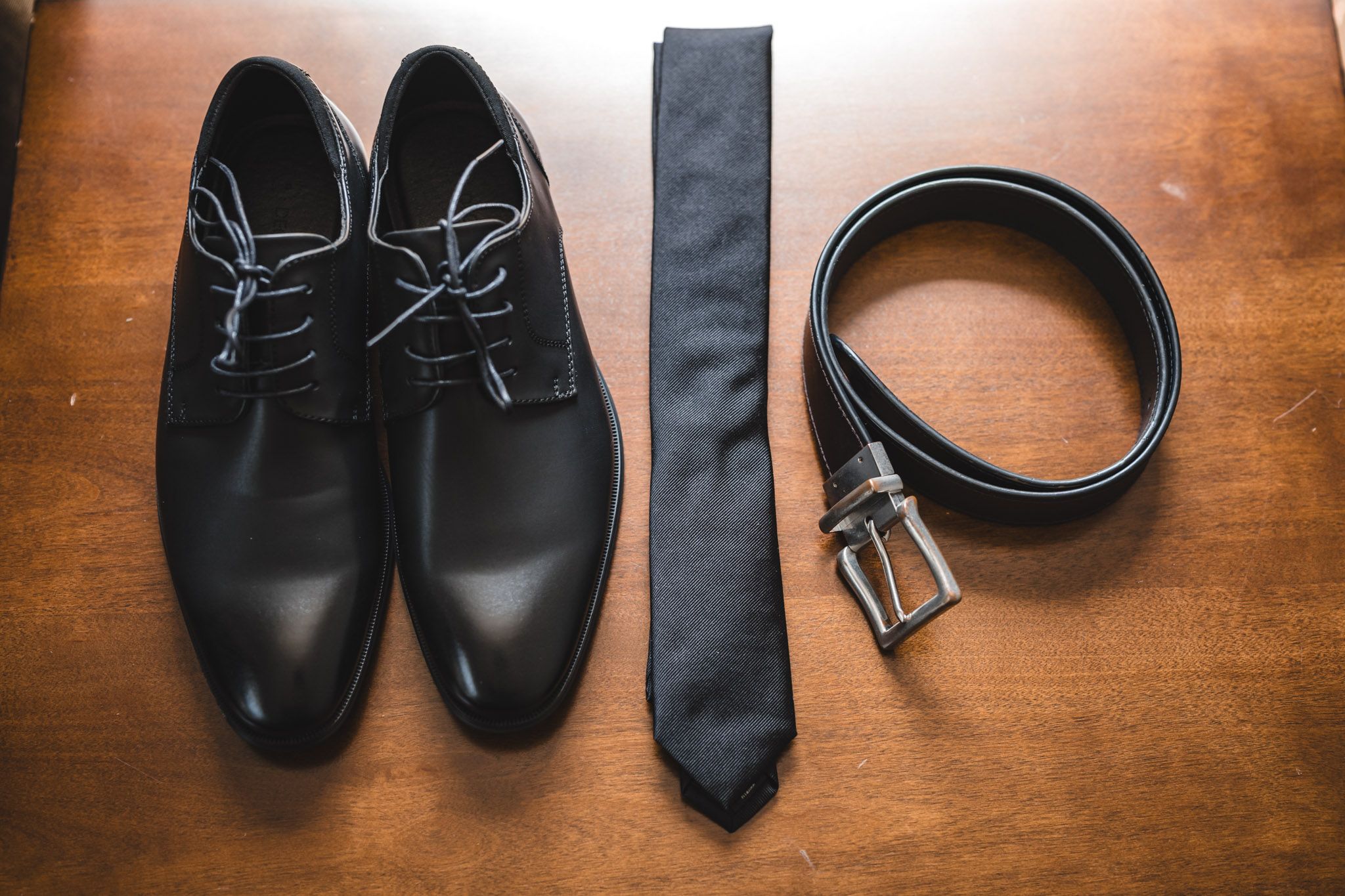 Groom's accessories including black dress shoes, tie, and belt arranged on wooden surface.