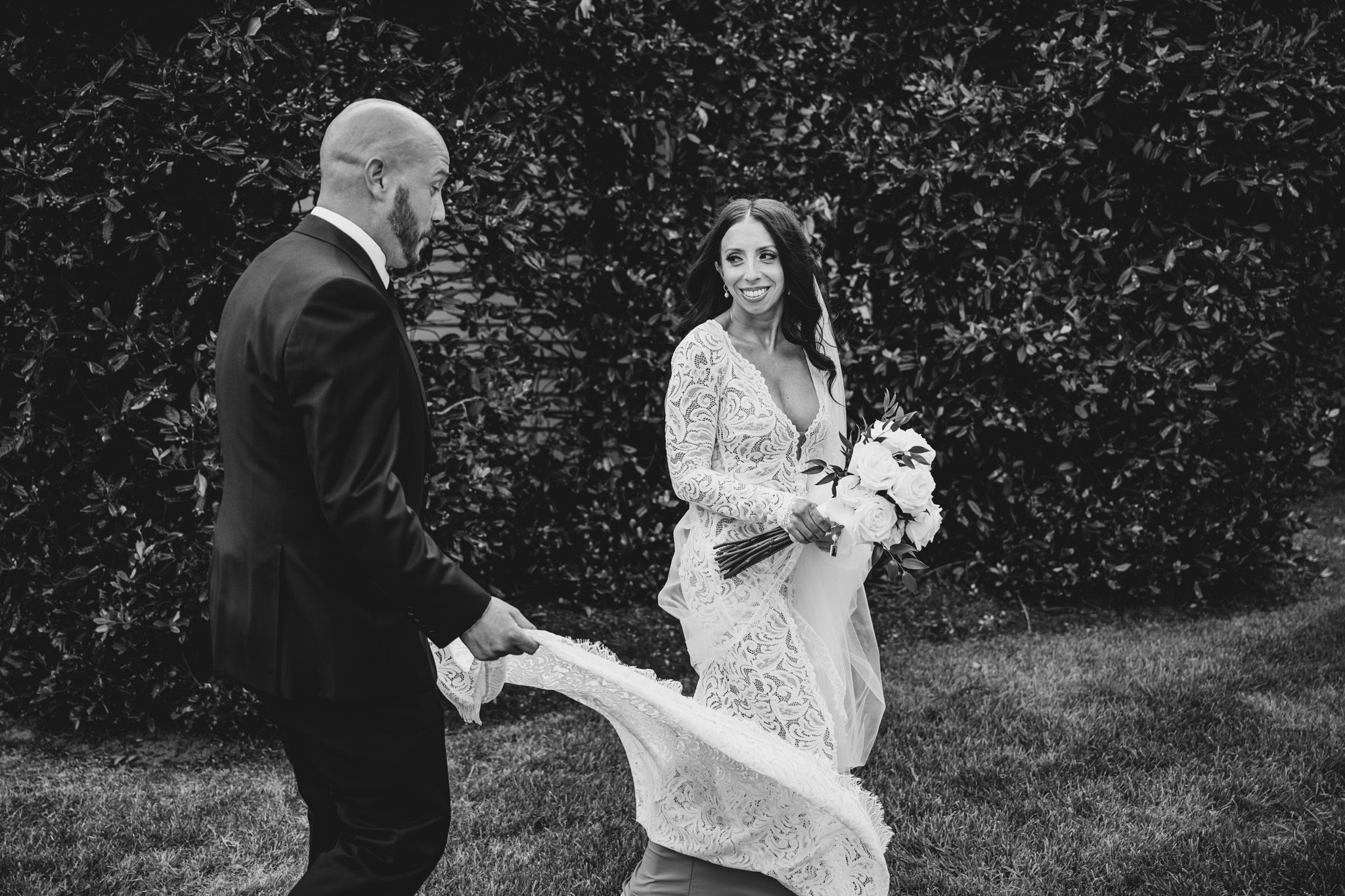 Playful wedding moment as breeze catches long white veil in garden setting.
