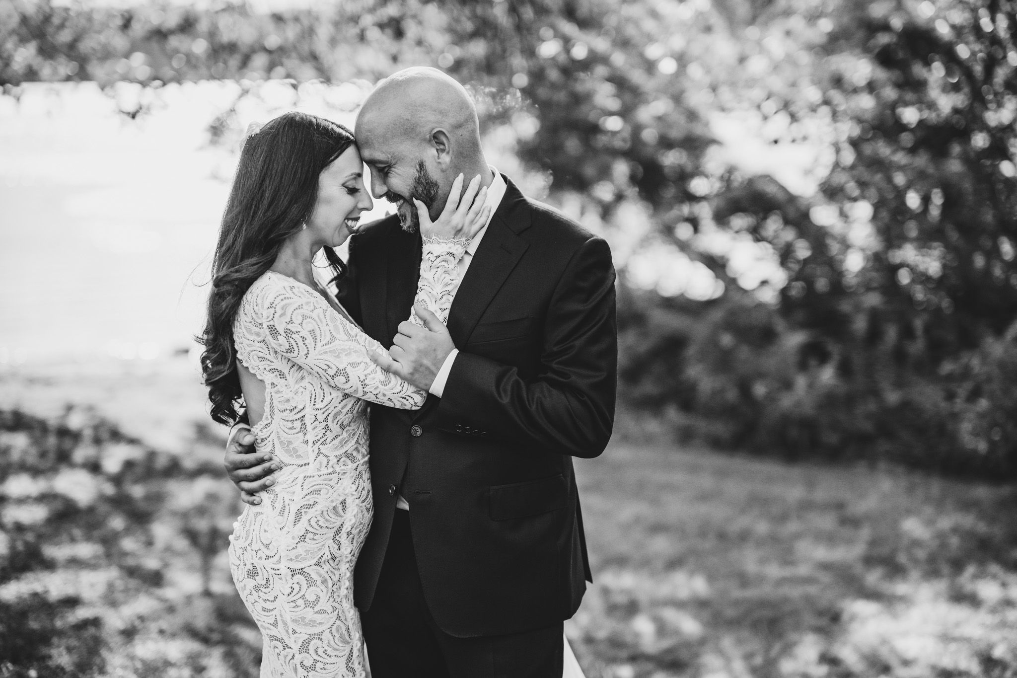 A romantic wedding couple dances together on a grassy lawn surrounded by trees.