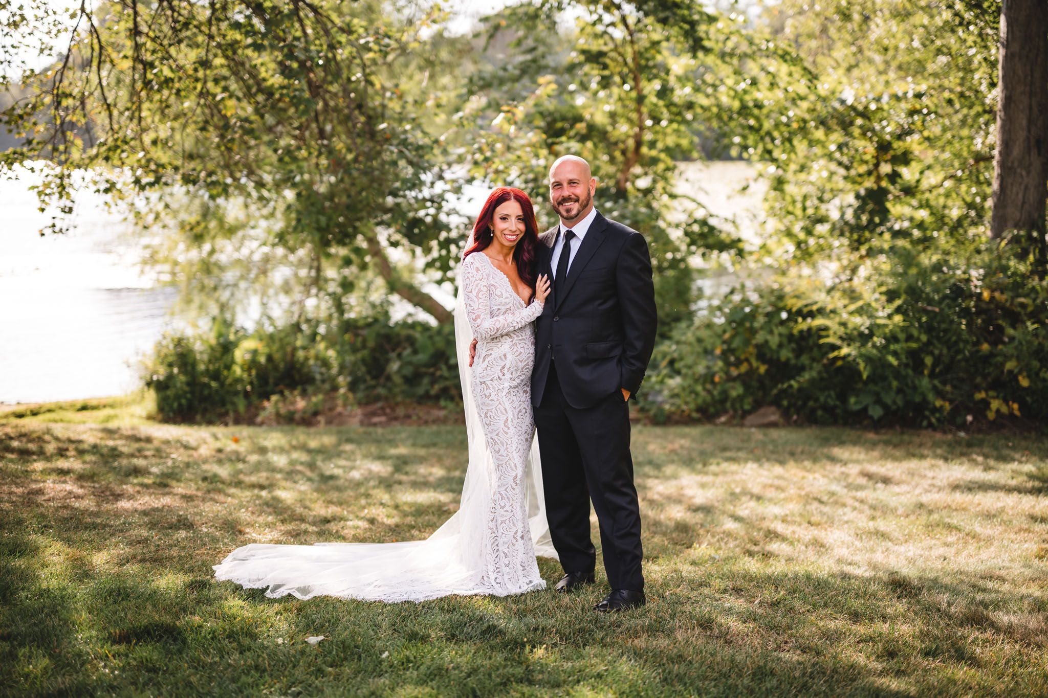 Wedding couple poses together on a sunny lawn with trees in the background.