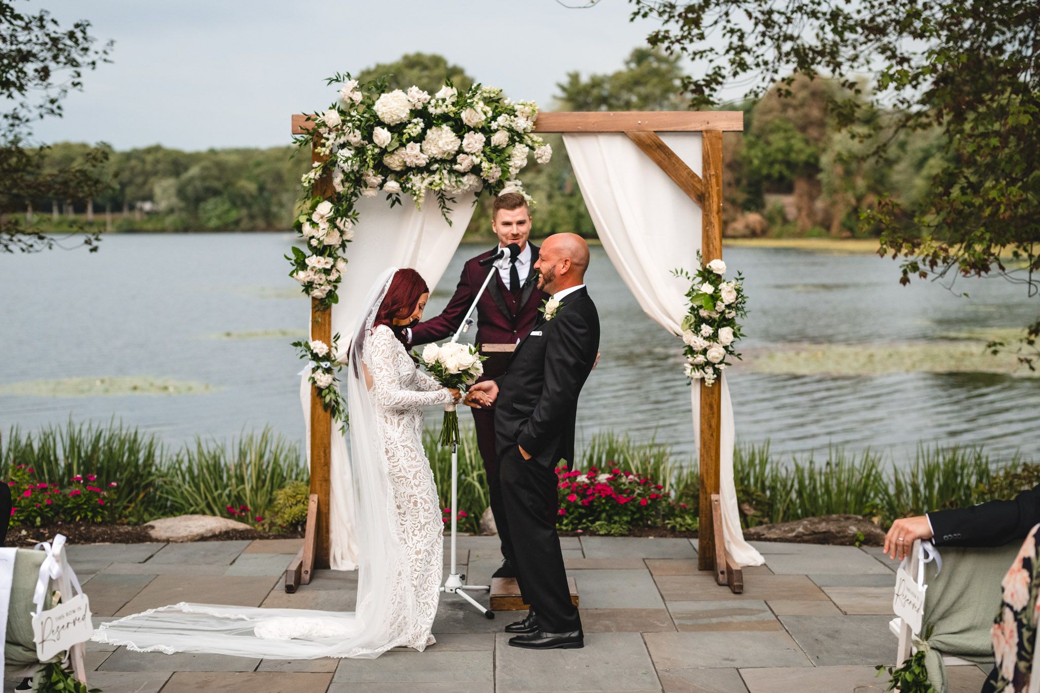 Outdoor wedding ceremony with wooden arch decorated with white flowers overlooking a lake.