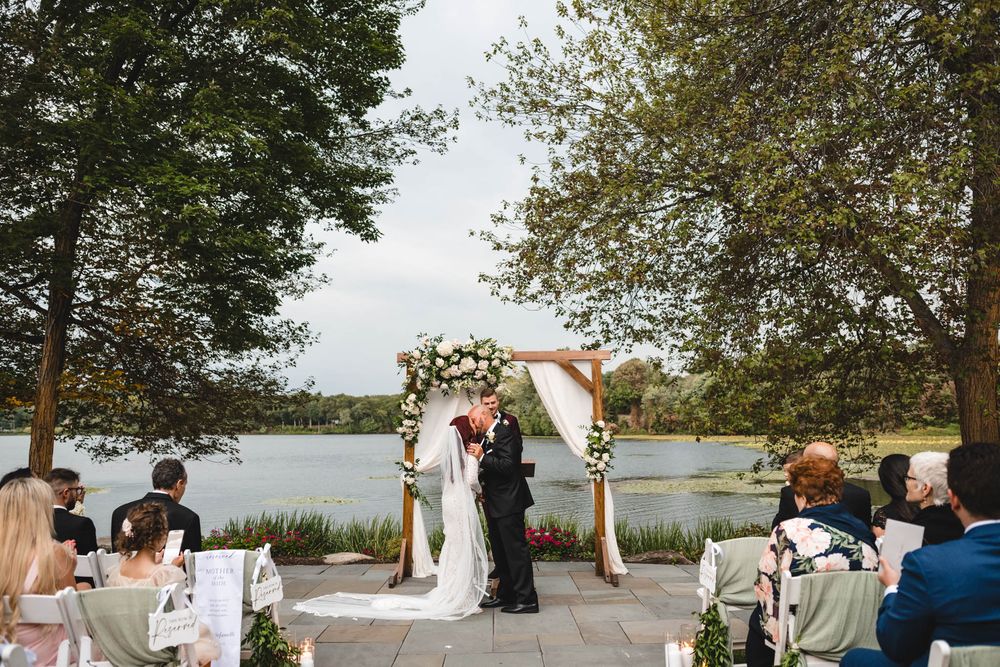Wide shot of lakeside wedding ceremony with floral arch and seated guests.