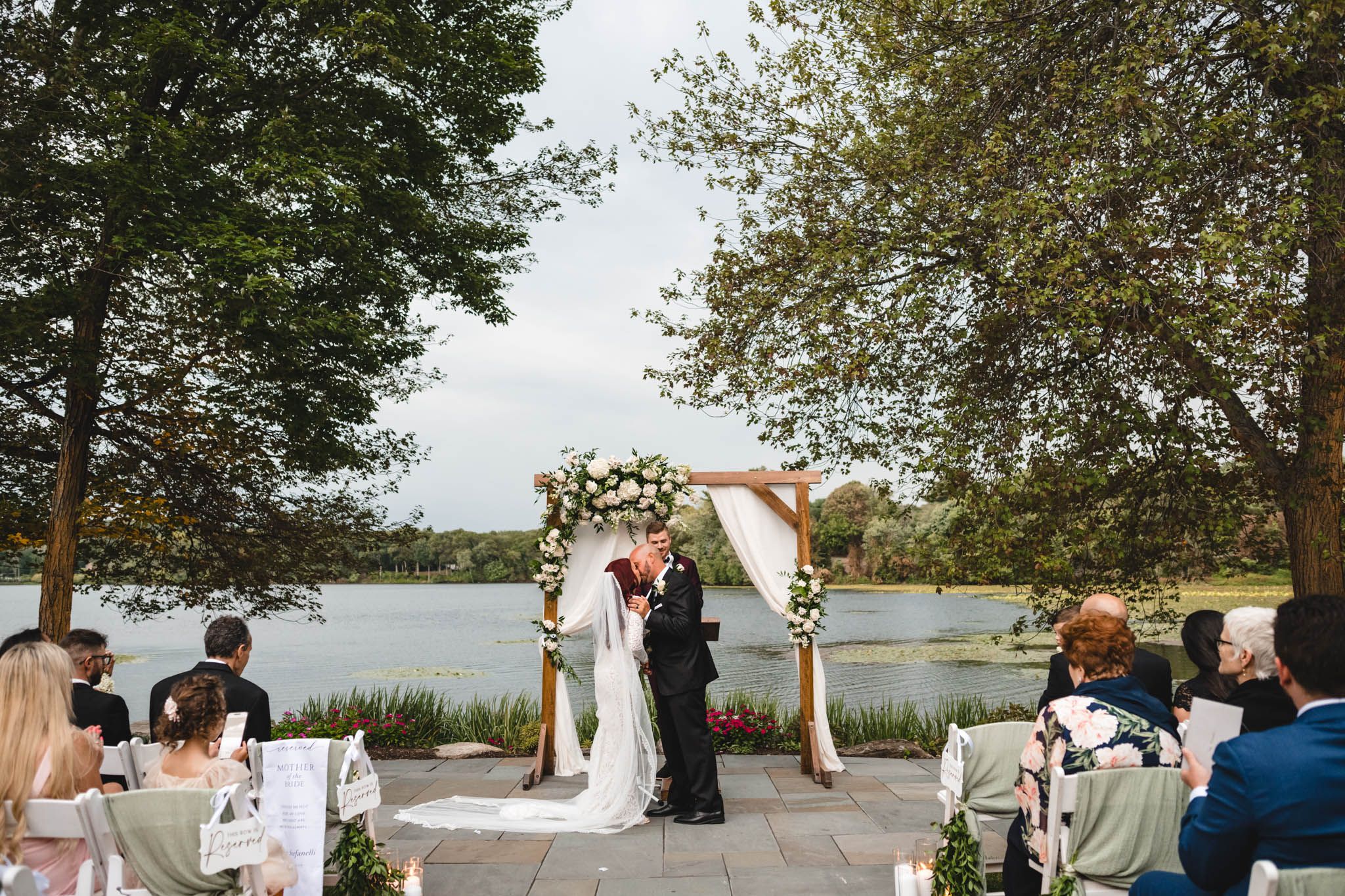 Wide shot of lakeside wedding ceremony with floral arch and seated guests.