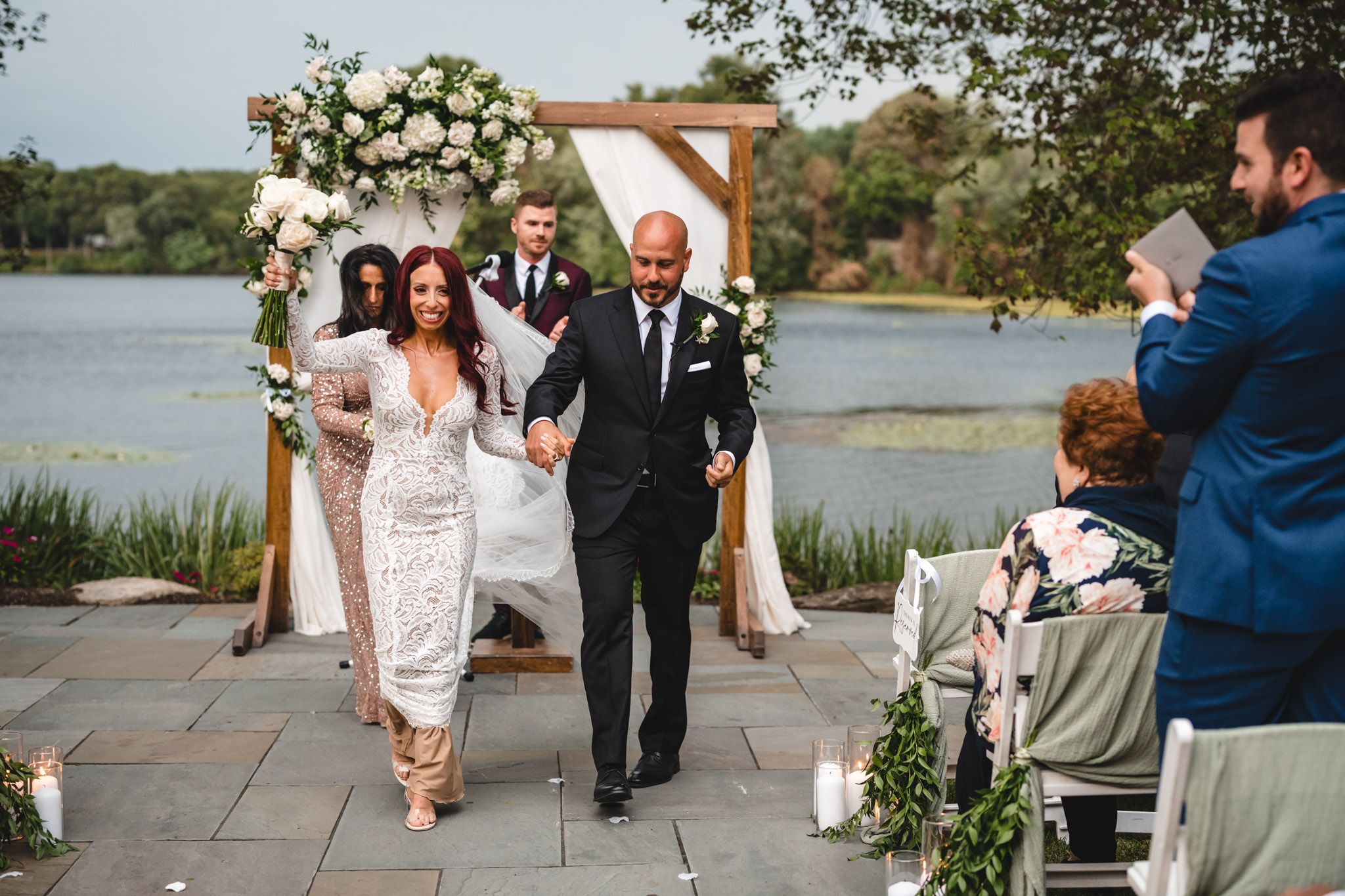 Wedding recessional on stone patio with floral arch and lake backdrop.