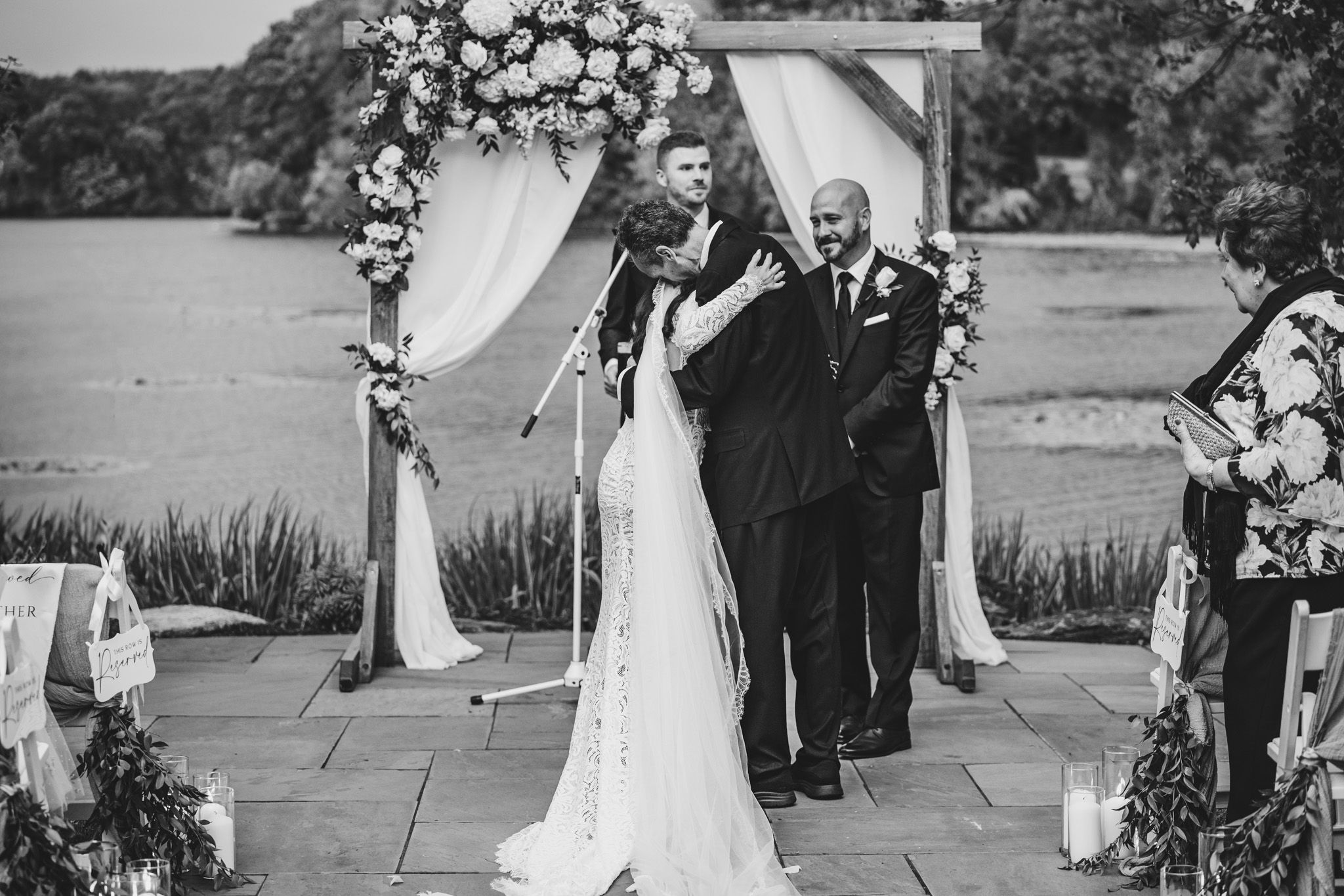 Black and white photo of outdoor wedding ceremony under floral arch by lakeside.