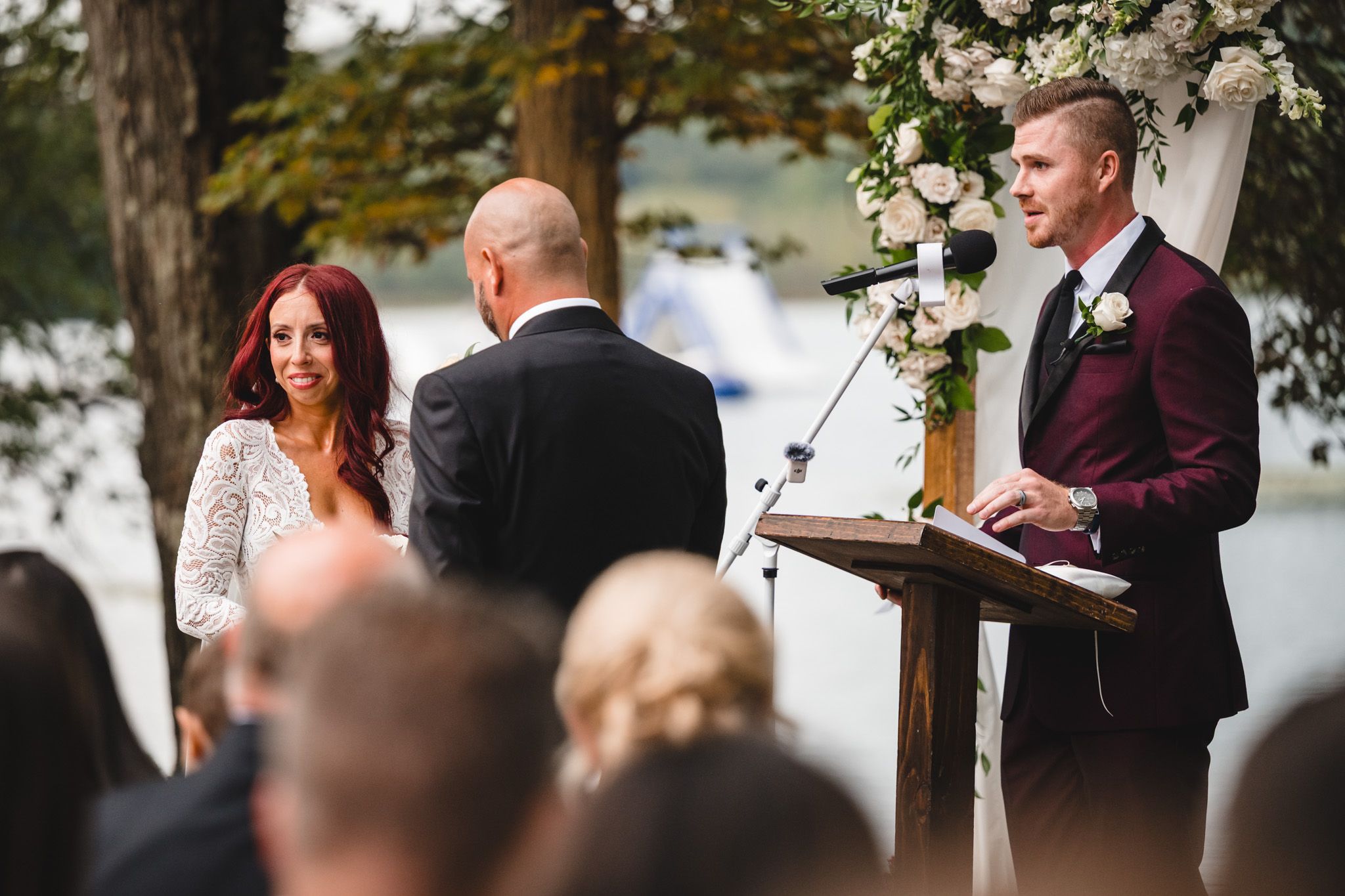 Outdoor lakeside wedding ceremony with floral arch and burgundy accents.