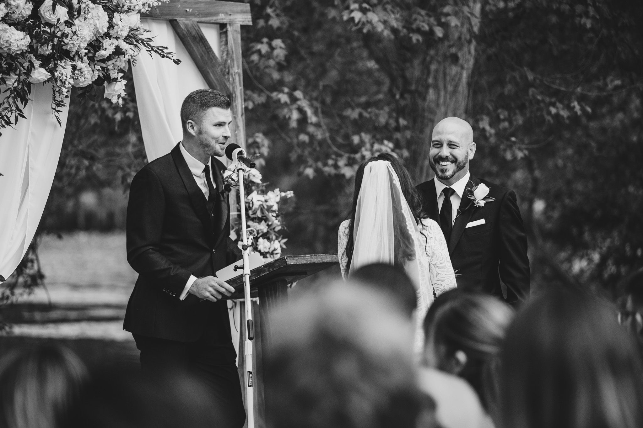 Black and white photo of wedding ceremony with speaker at microphone outdoors.