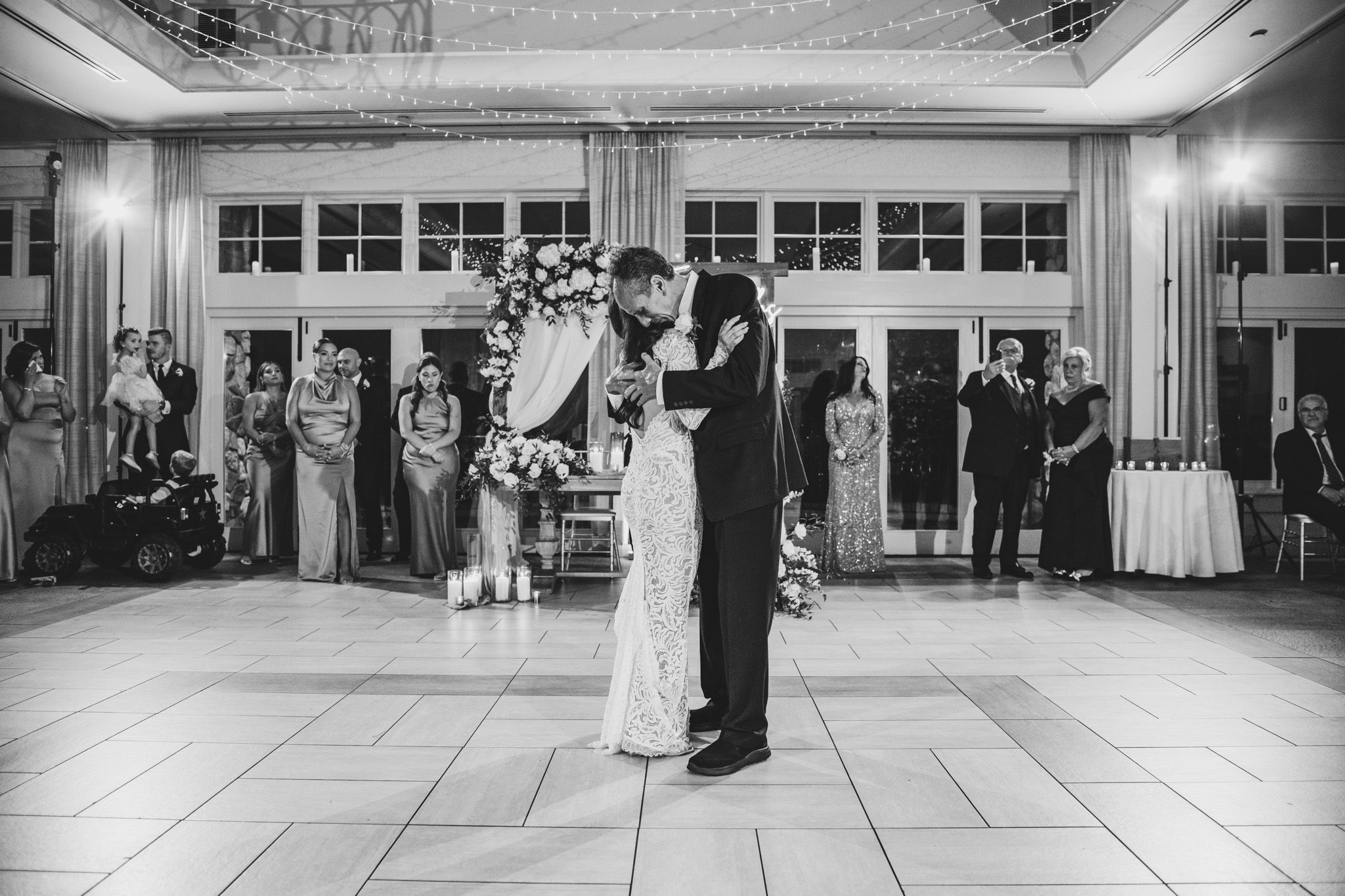 A black and white photograph of a wedding couple dancing while guests look on in an elegant venue.