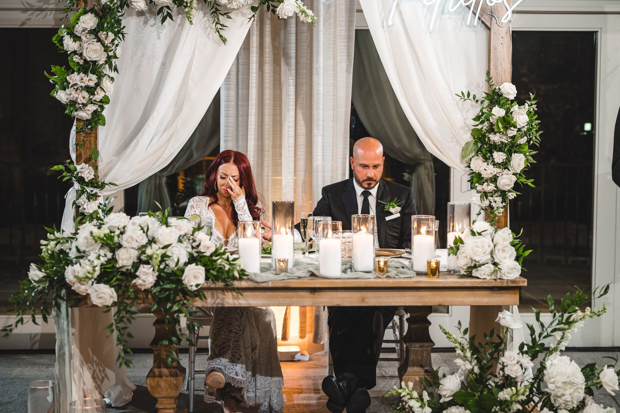 Wedding reception table decorated with white floral arrangements, candles, and elegant white draping.
