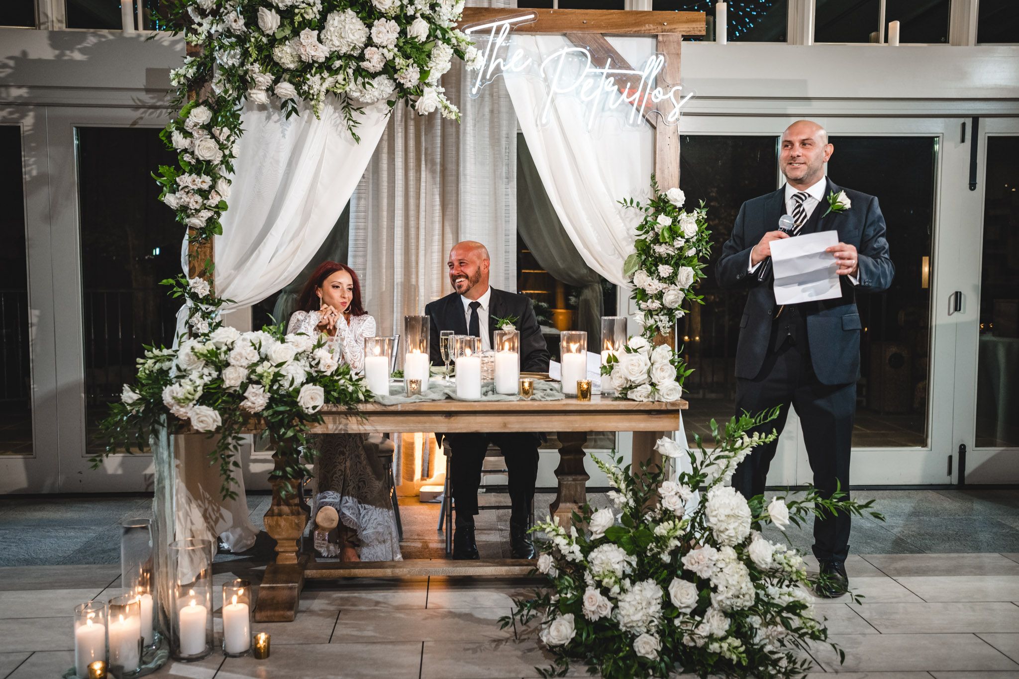 Wedding reception setup with elegant white floral arrangements and candles at the head table.