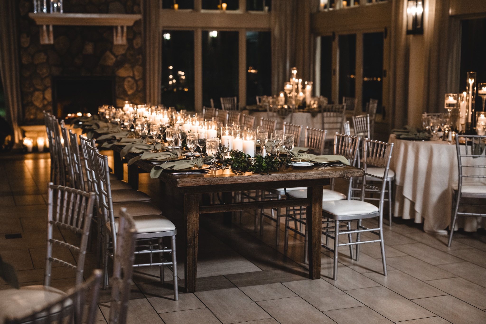 Elegant wedding reception table setup with rustic wooden tables, silver chiavari chairs and glowing candlelight.