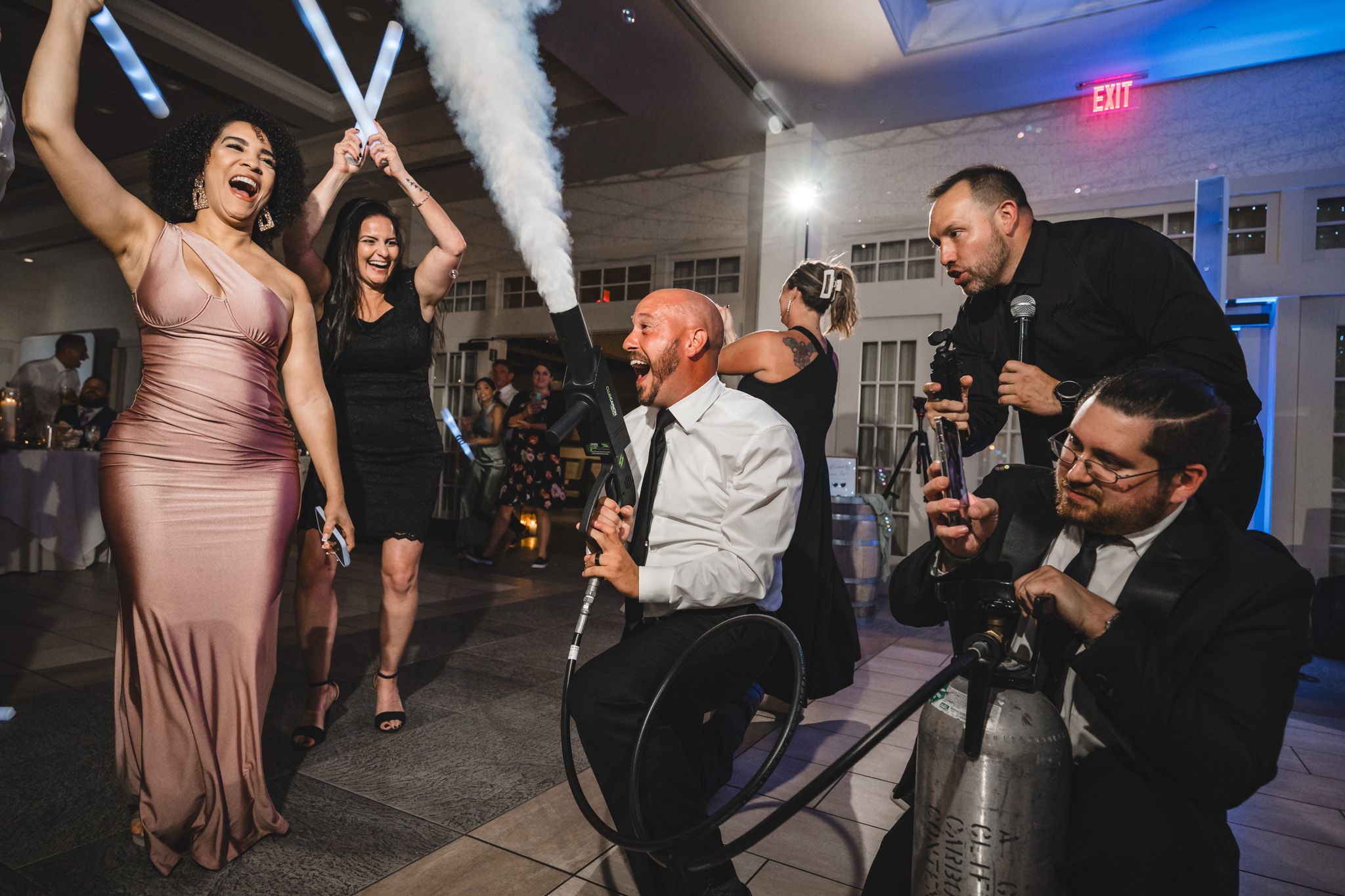 Party guests dance and celebrate with light-up sabers under string lights at an indoor wedding reception.