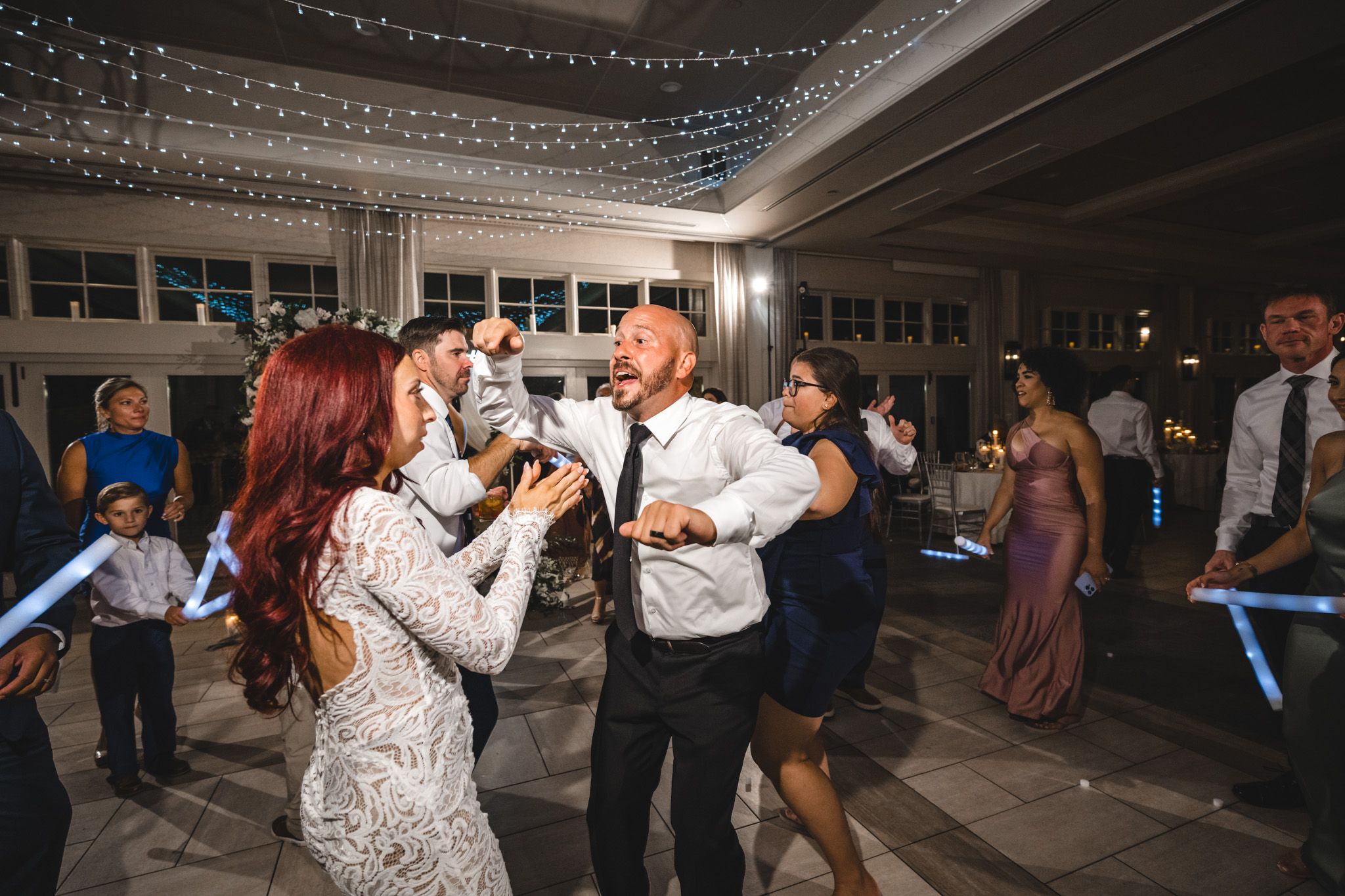 Wedding party guests dance with illuminated sabers under a canopy of string lights.
