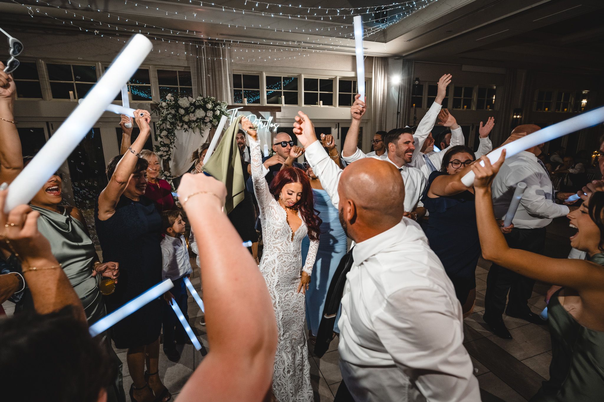 Guests hold glowing light sabers high during a nighttime wedding celebration under twinkling lights.