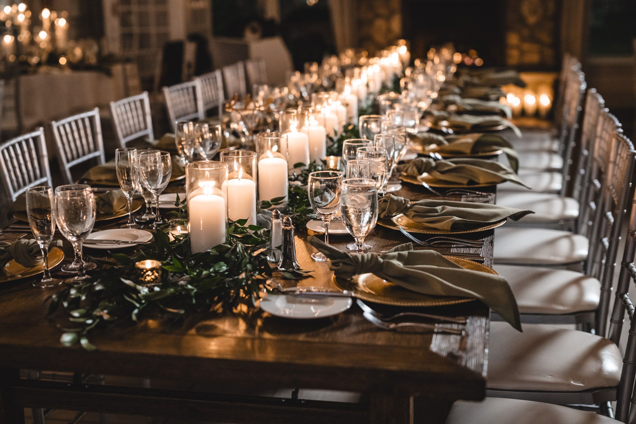 Long banquet table decorated with white candles and greenery garland for an elegant wedding reception.