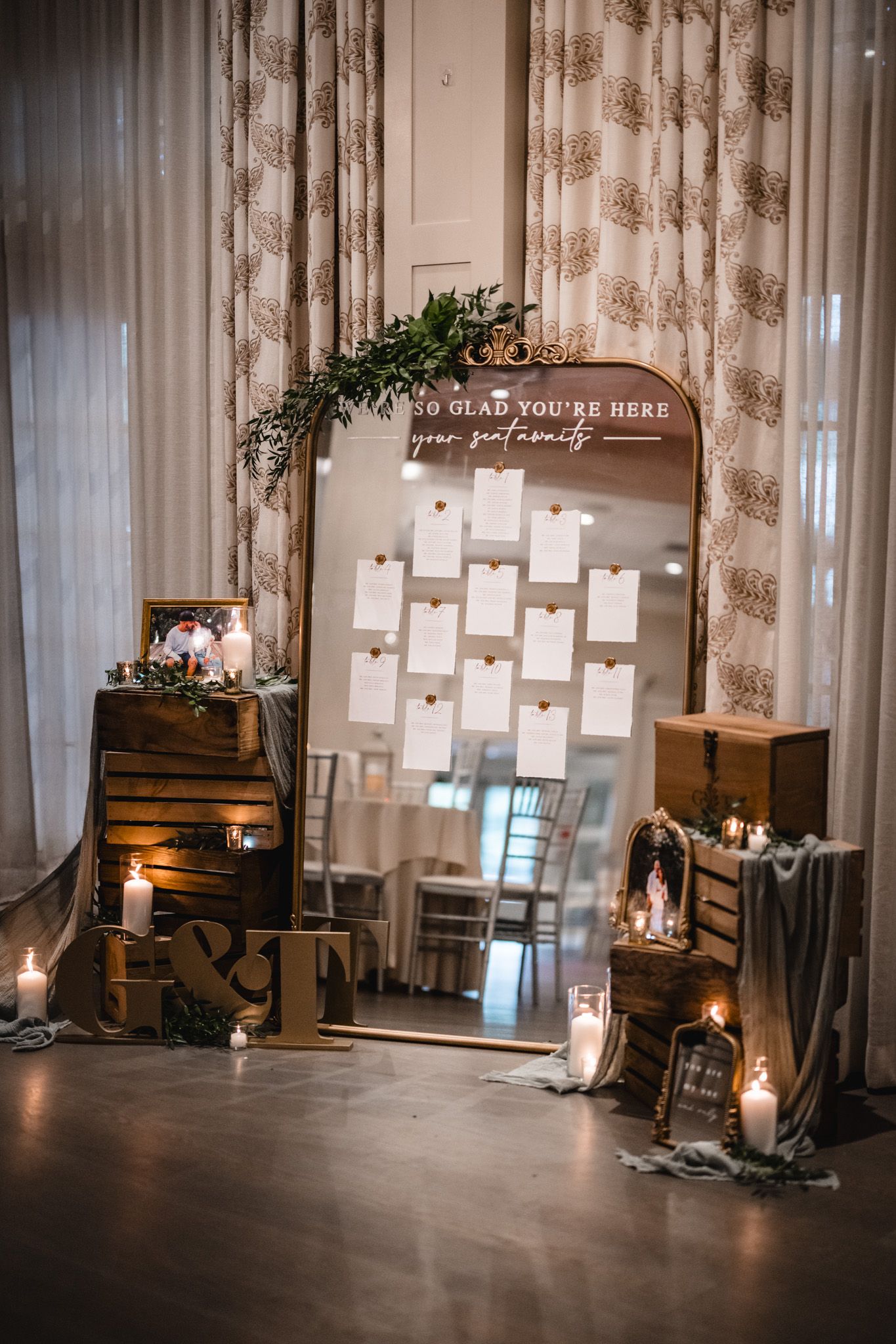 Wedding seating chart display with wooden crates, candles and greenery against floral curtains.