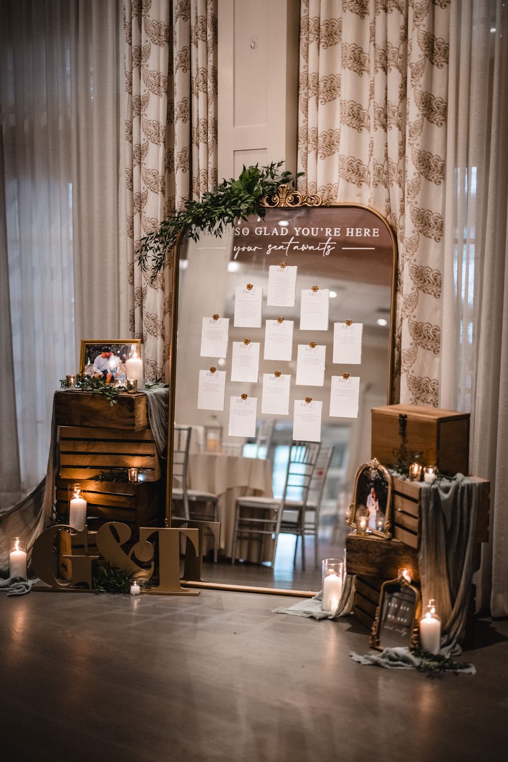 Wedding seating chart display with wooden crates, candles and greenery against floral curtains.