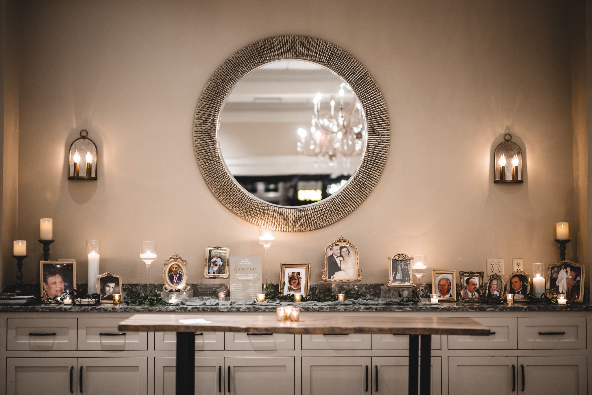 Elegant wedding photo display on buffet cabinet beneath round mirror with wall sconces.