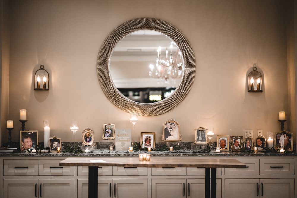 Elegant wedding photo display on buffet cabinet beneath round mirror with wall sconces.