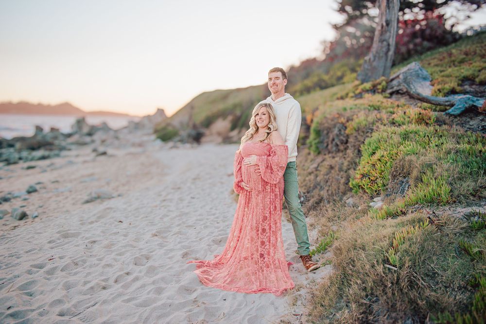 Pregnant woman in flowing pink lace gown poses with partner on sandy beach at golden hour.