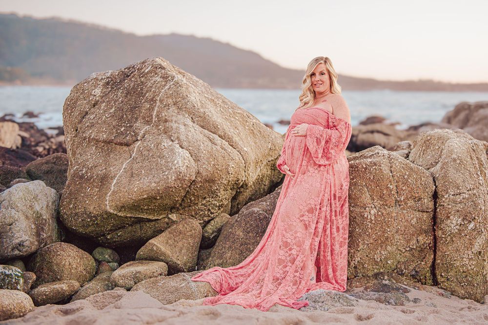 Pregnant woman in pink lace maternity gown posing on rocky beach at sunset.