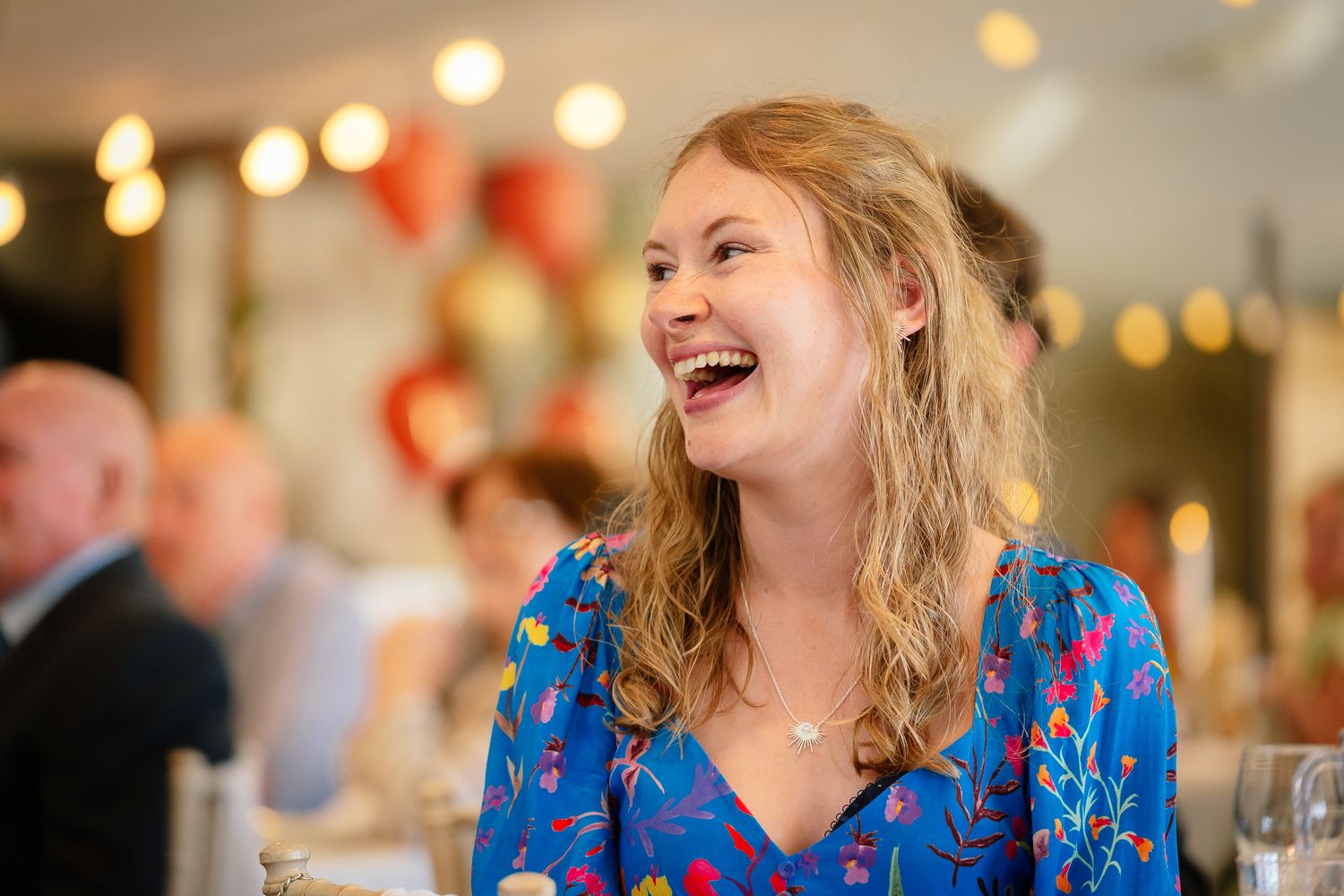 A person in a bright blue patterned dress laughs joyfully at an indoor event with glowing lights in the background.